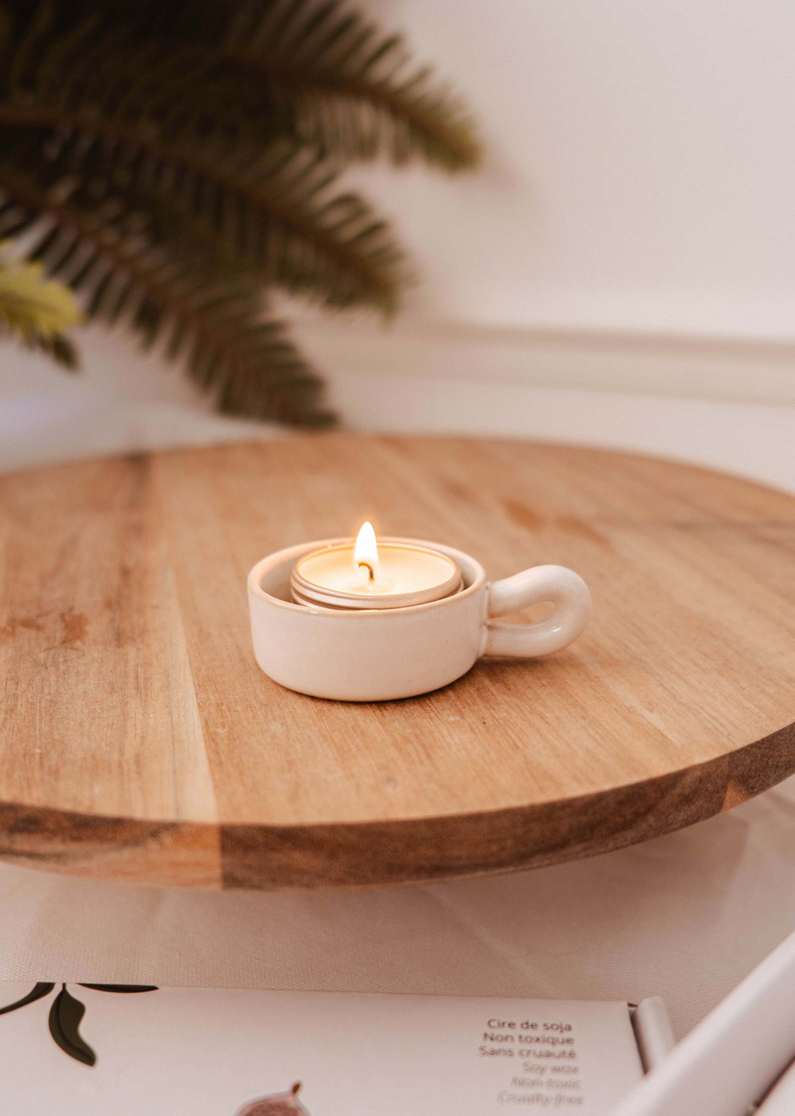 A lit tealight sits in the Mimi & August Tealight Holder, a small white ceramic cup on a wooden surface, bringing charm to rustic decor. A leafy plant adds a natural touch in the background.