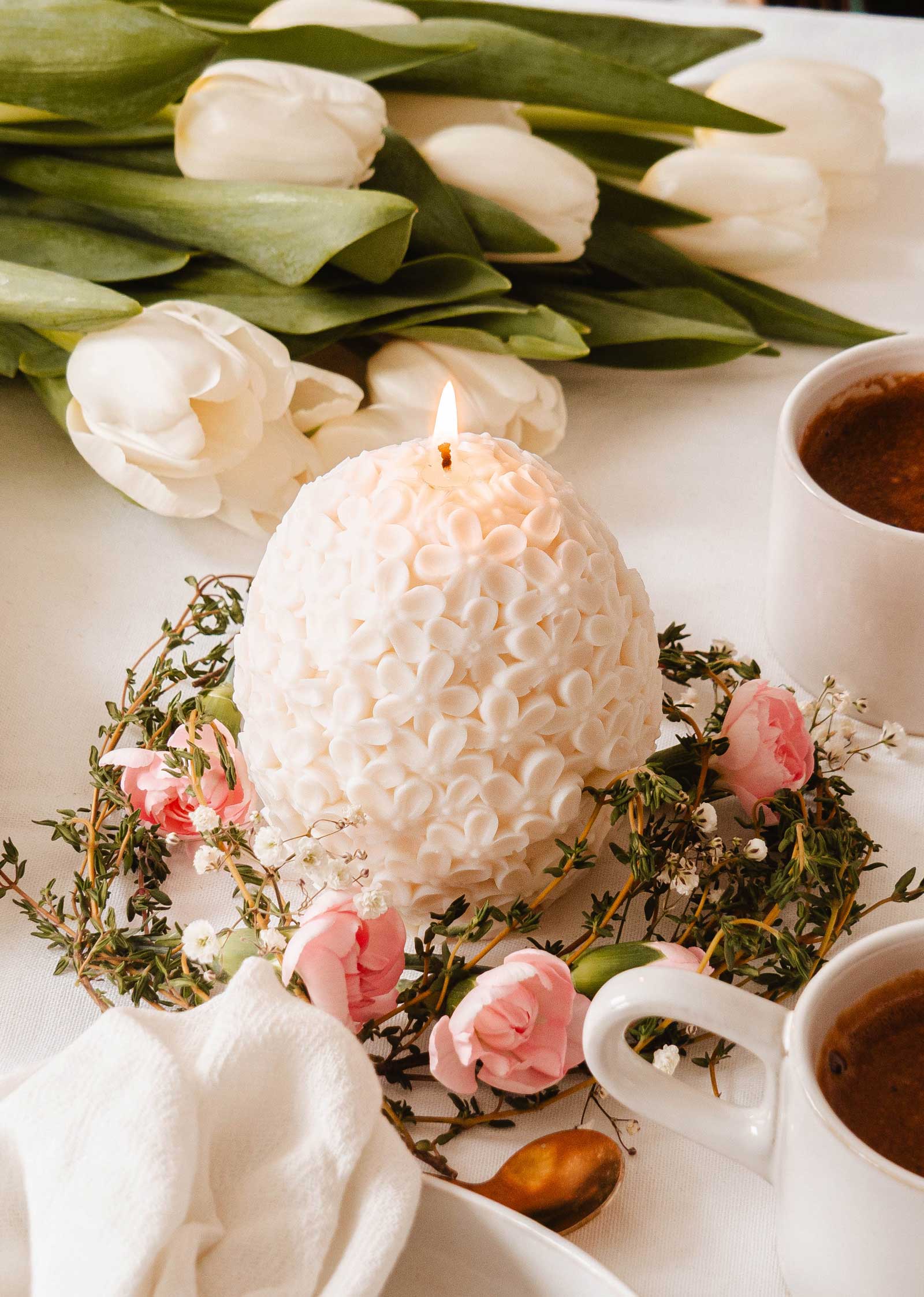 A Floral Egg Decorative Candle by Mimi & August, with exquisite pink and white flowers encircling its glowing floral patterns, is set amid two cups of coffee and a bouquet of white tulips on a sophisticated white table.