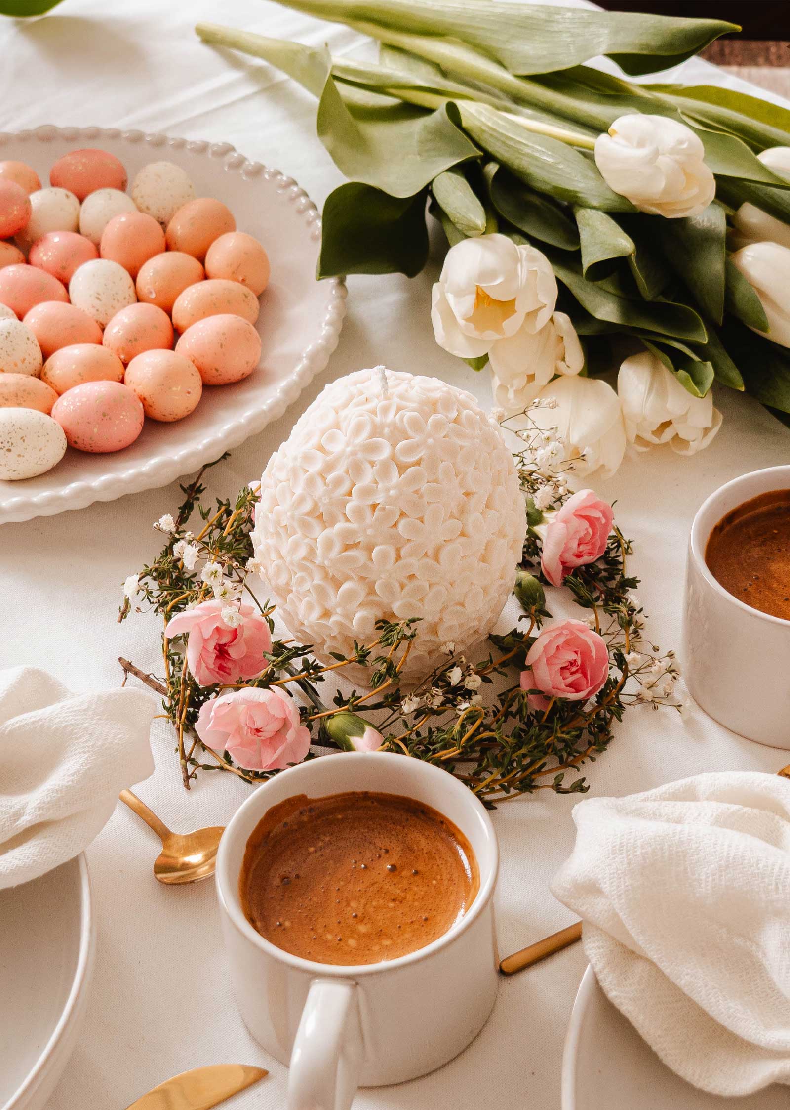 The table is adorned with The Floral Egg decorative candle by Mimi & August as a centerpiece, surrounded by pink roses, pastel eggs, white tulips, and paired with two coffee mugs—creating an exquisite and elegant setting.