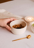 A hand holds a white bowl filled with granola on a white tablecloth, accompanied by Mimi & August's The Glow Candle and a small gold spoon.