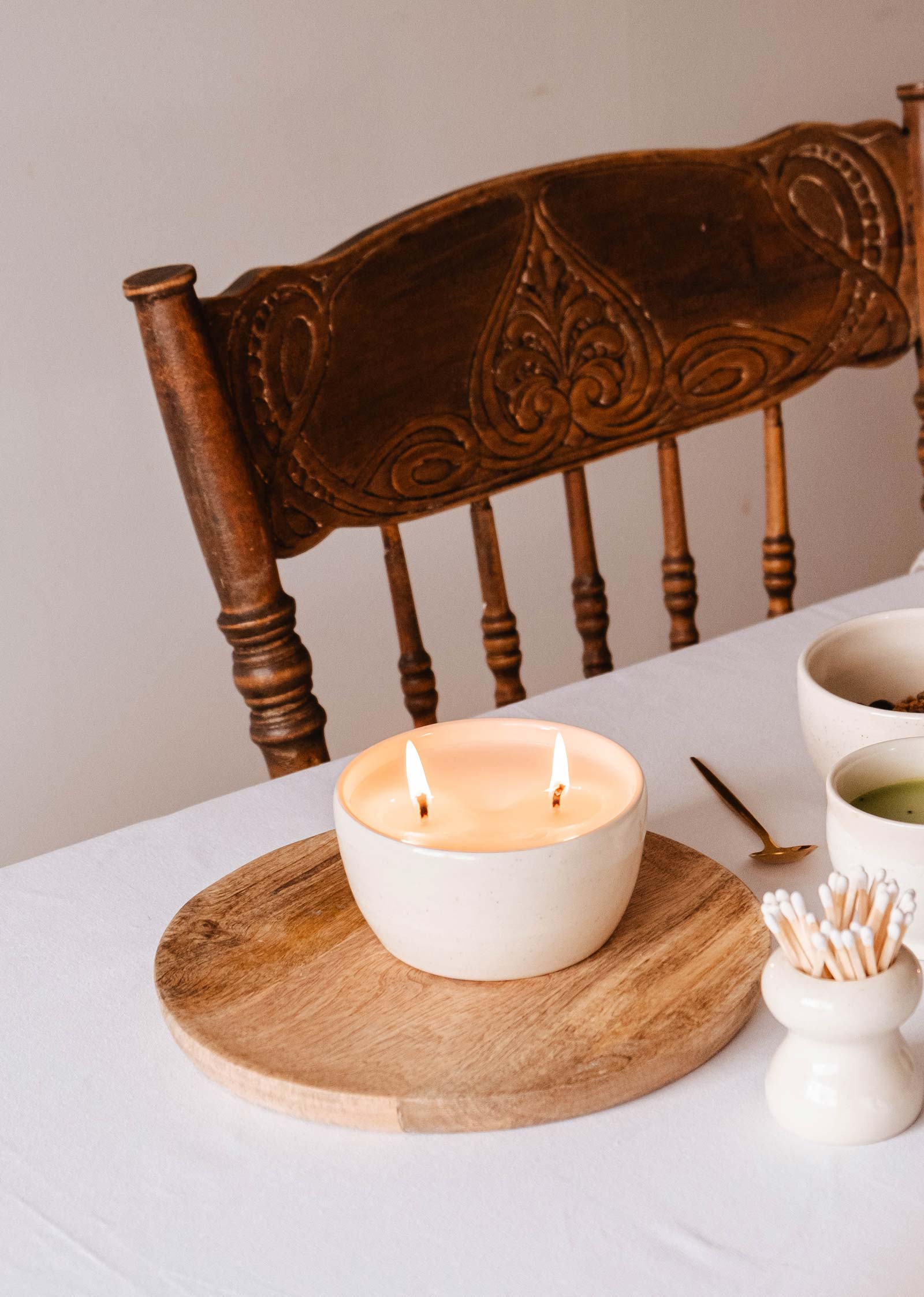 The Glow Candle by Mimi & August, a handcrafted ceramic with two wicks in a white bowl on a wooden platter, creates an inviting glow on a white table. Its enchanting scents fill the air alongside a wooden chair and bowls with matches and greenery, embodying Diane Brouillette's serene artistry.