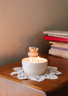 The Glow Candle by Mimi & August, a handcrafted ceramic piece, sits in a white bowl on a doily, casting enchanting scents as it rests next to a stack of books on the wooden table.