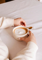 Une personne tient une tasse de café sur une table blanche avec une cuillère dorée et la bougie lumineuse de Mimi & August, fabriquée à la main par Diane Brouillette, infusant l'air de senteurs uniques.