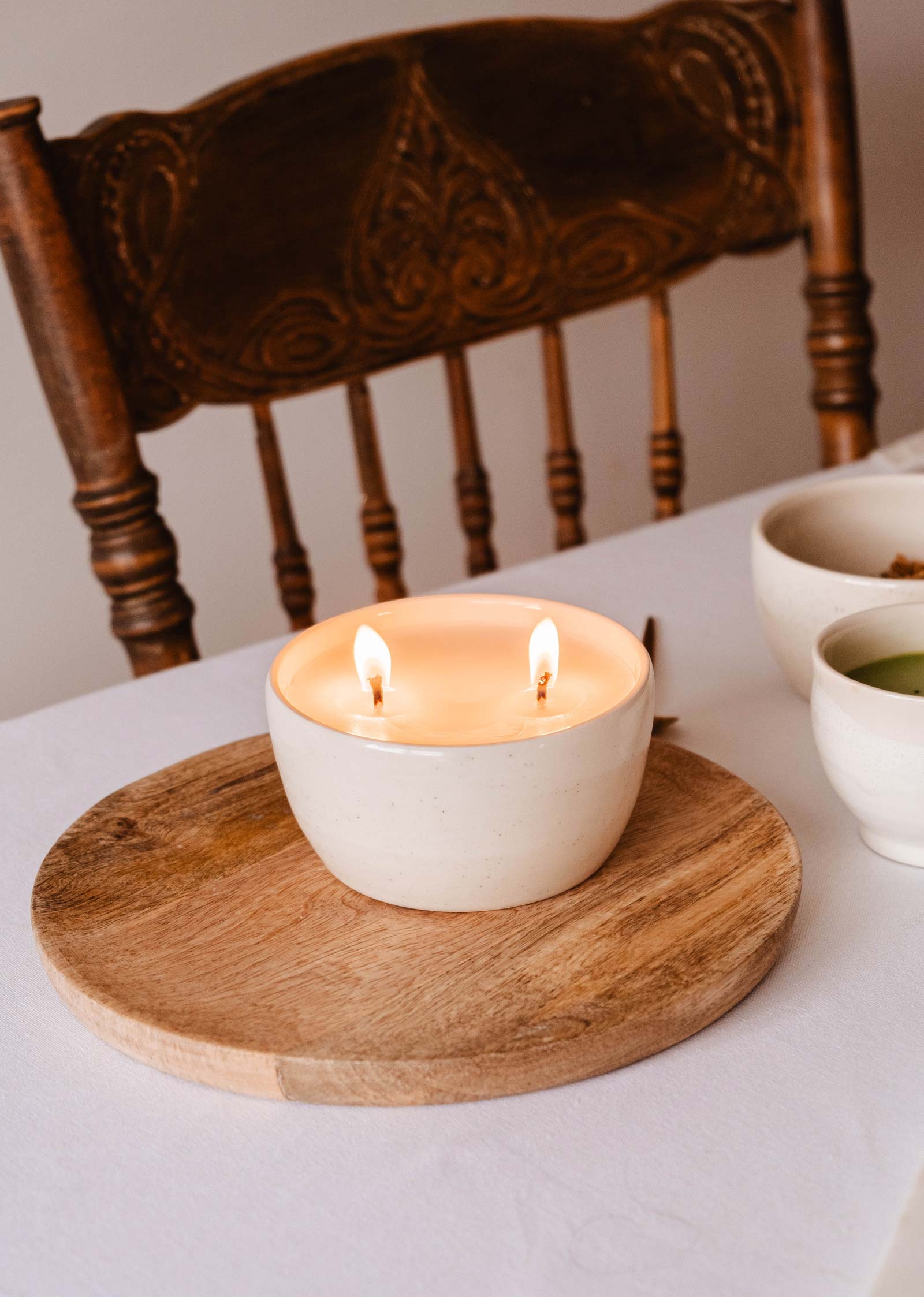 The Glow Candle by Mimi & August, featuring two unique flames in a white ceramic holder on a wooden board, flickers softly and adds charm to the table. In the background, a wooden chair completes this cozy scene.