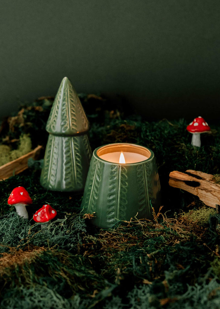 The Green Fir Tree by Mimi & August is a candle housed in a tree-shaped holder, releasing the comforting scent of fir trees. It sits on a bed of moss and is adorned with tiny red mushrooms and wooden accents, all set against a moody dark backdrop.