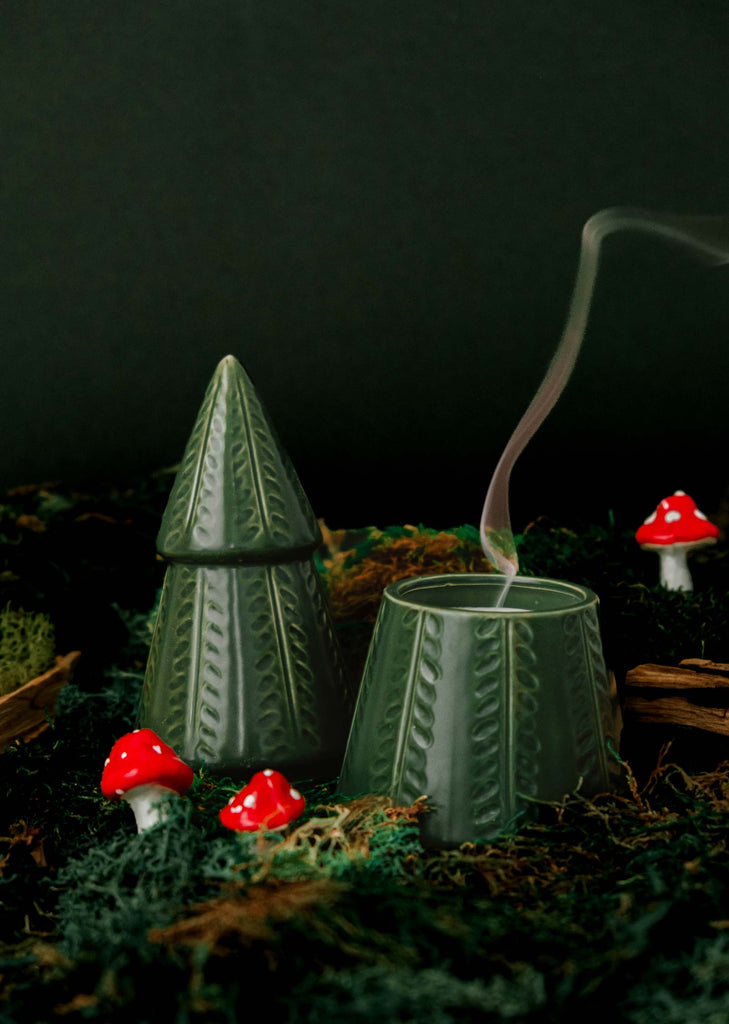 Introducing "The Green Fir Tree" by Mimi & August, a charming green fir tree candle holder designed in the shape of a pine. It emits a gentle wisp of smoke and is beautifully accentuated by small red mushroom figurines resting on a bed of moss, capturing the enchanting Christmas fragrance.