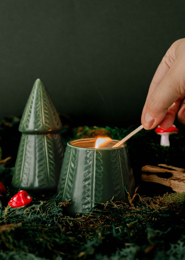 A hand lights "The Green Fir Tree" Christmas candle by Mimi & August, featuring a design that includes moss, small mushrooms, and rustic decor.