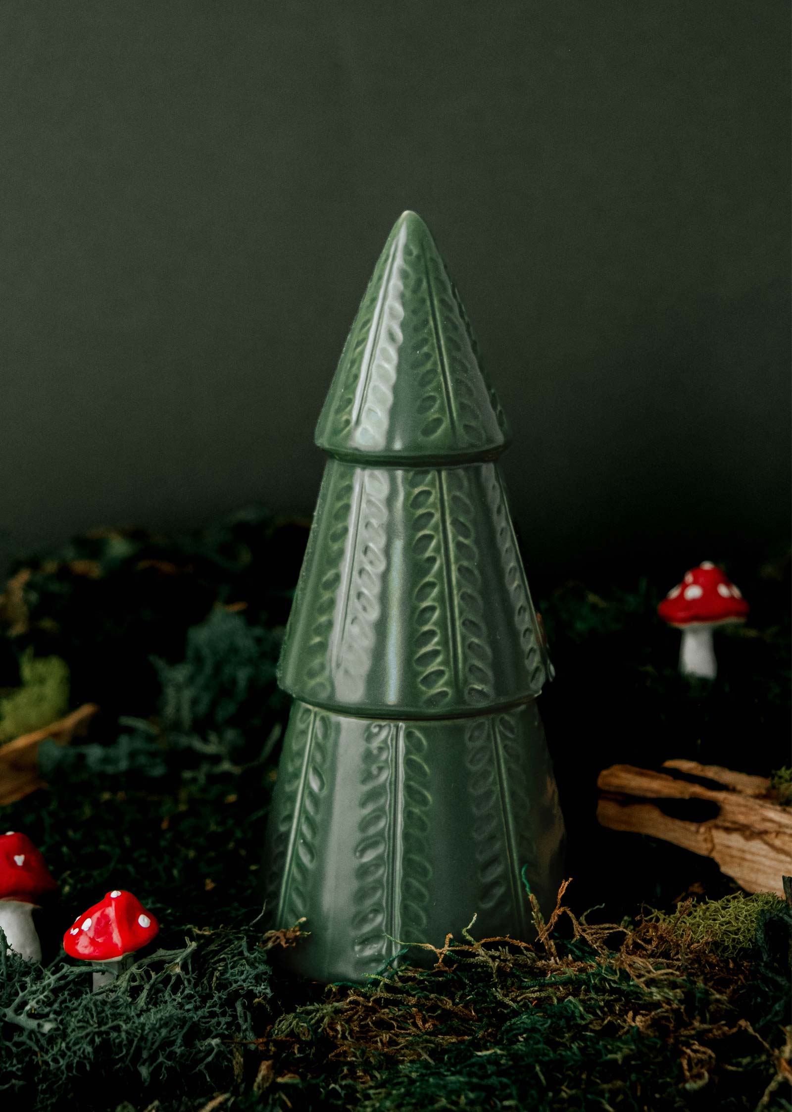 Un arbre vert en céramique orne une surface moussue, complété par de petites figurines de champignons rouges et blancs. À proximité, le parfum apaisant de la bougie "Le sapin vert" de Mimi & August ajoute une touche invitante à cette scène enchanteresse.