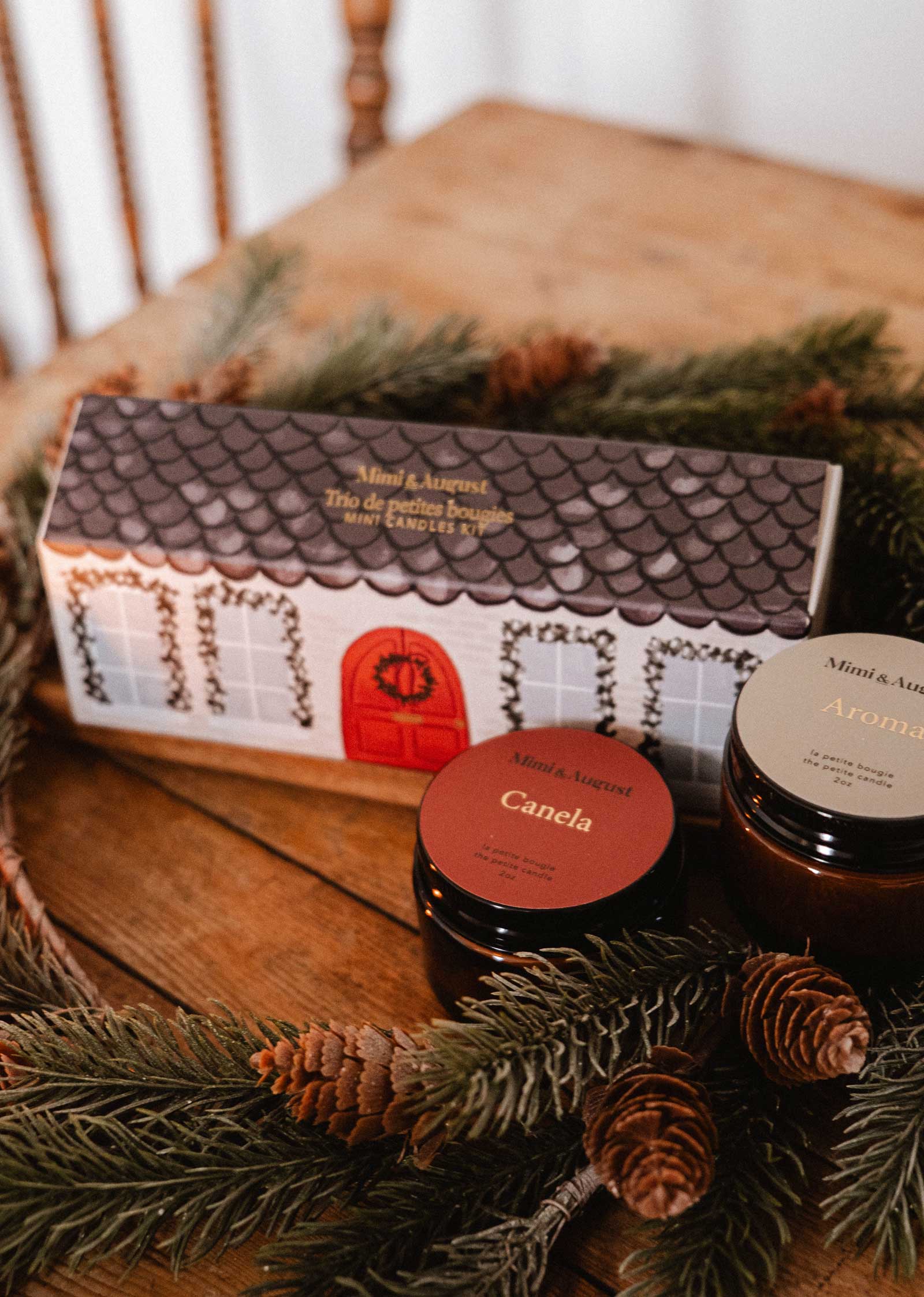 The Little Christmas Box by Mimi & August features two labeled, hand-poured candles in jars and a house-shaped box, all arranged with pine branches and pinecones for a festive touch on a wooden table.