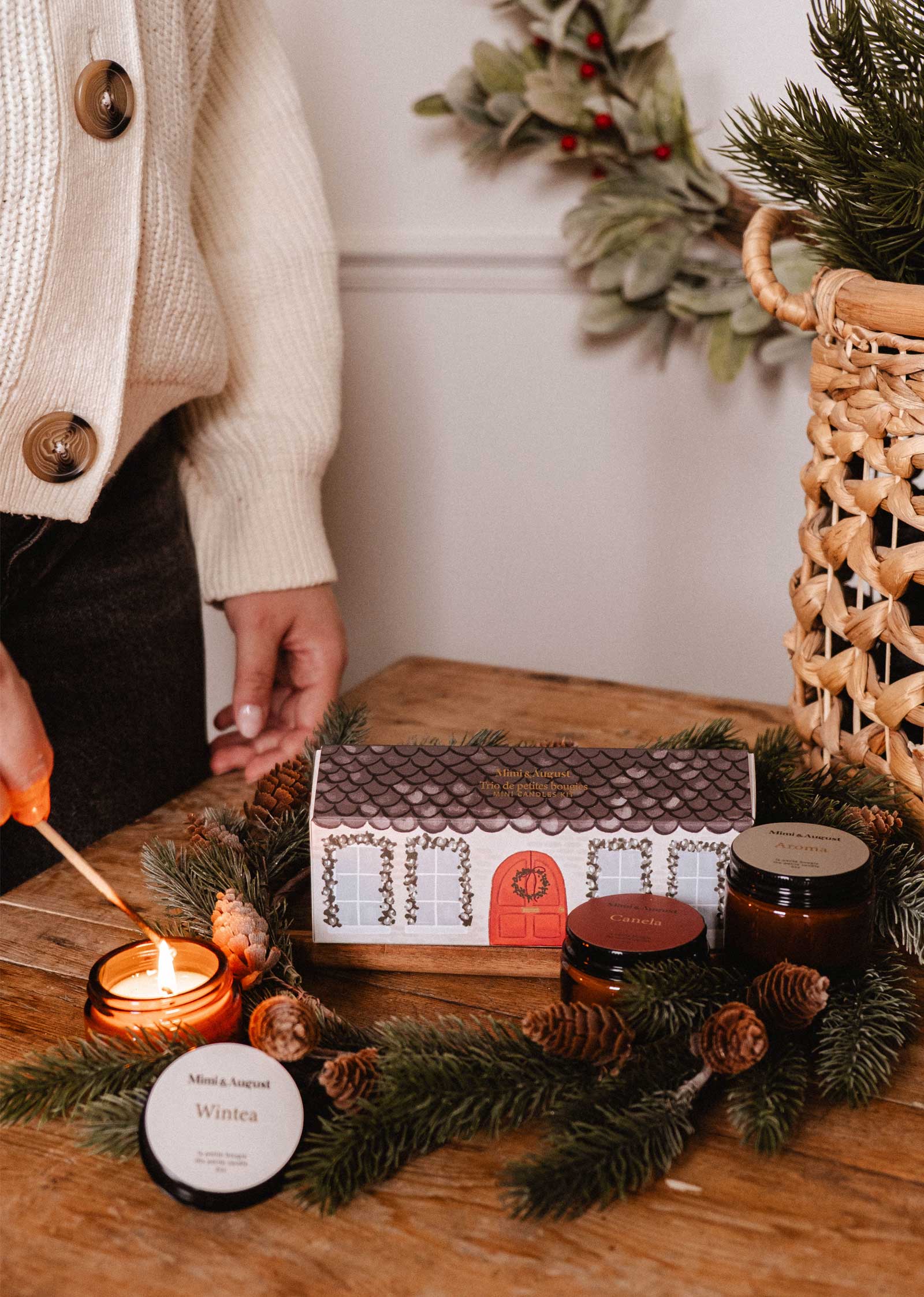 A person lights Mimi & August's The Little Christmas Box candle on a wooden table adorned with pine branches, pinecones, jars, and a small house-shaped box—a cozy setting to showcase this festive holiday scented candle.