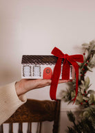 A hand holds Mimi & August's The Little Christmas Box—a house-shaped gift with a red door and big red ribbon, set against festive greenery—perfect for gifting holiday candles.