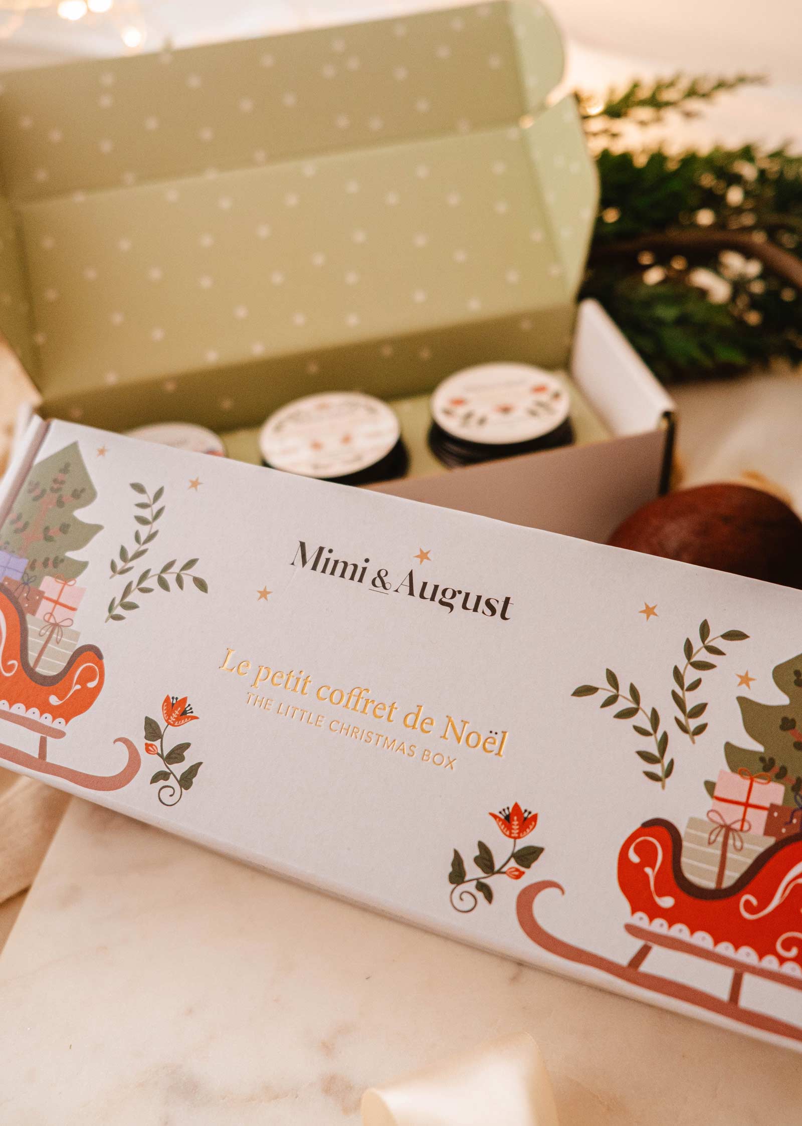 Un coffret festif intitulé "The Little Christmas Box" de Mimi & August, avec un design sur le thème de Noël et partiellement ouvert pour présenter des parfums de vacances et de délicieuses bougies de Noël.