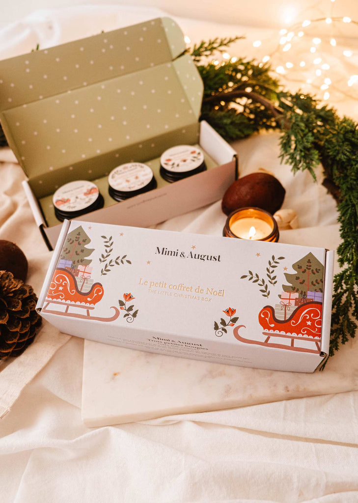 The Little Christmas Box by Mimi & August showcases three small jars infused with holiday scents, accompanied by greenery, a lit candle, and festive packaging perfect for the season's decor.