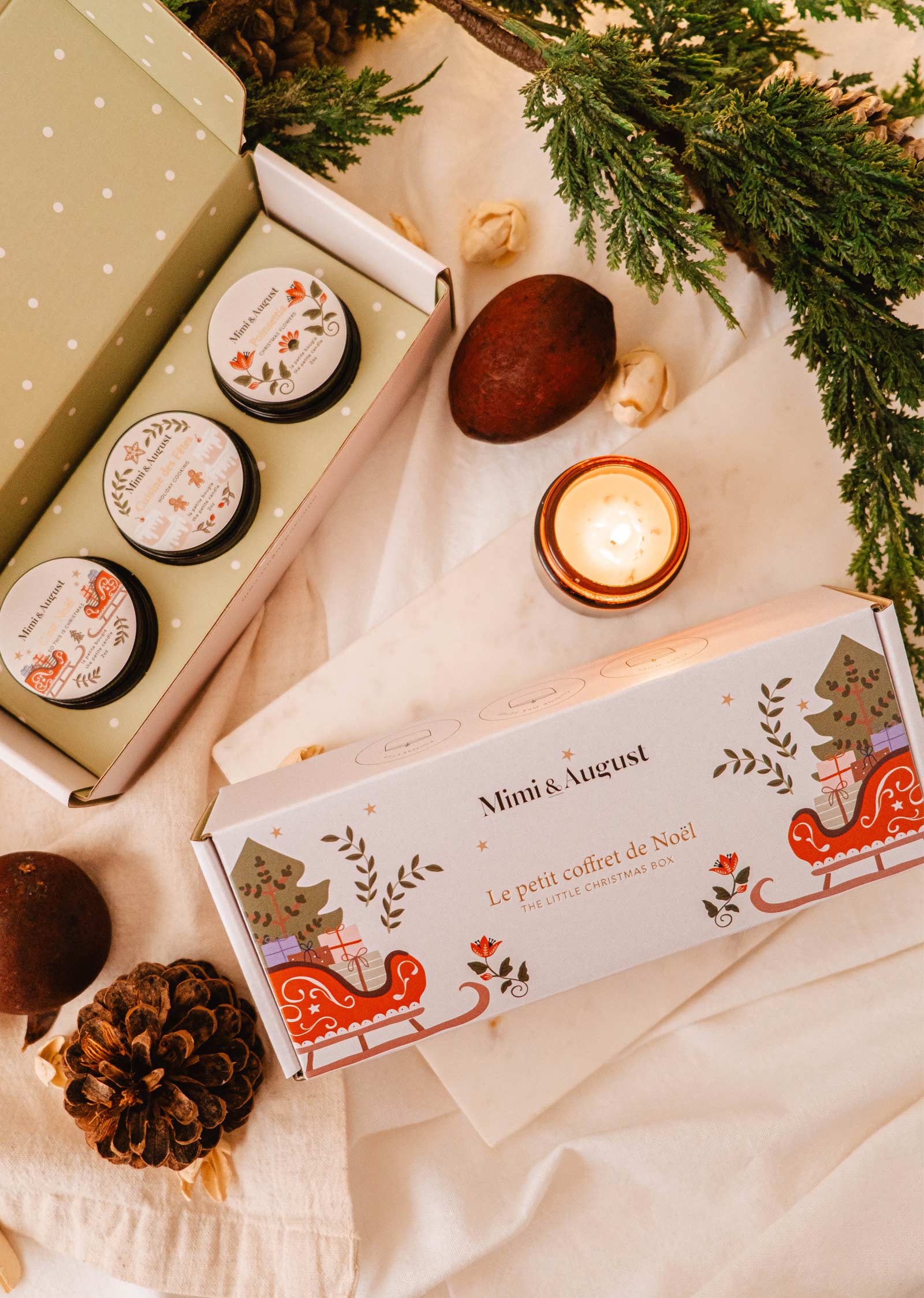 La Petite Boîte de Noël de Mimi & August est un coffret de Noël aux senteurs saisonnières et enchanteresses, avec des bougies de Noël dans un emballage festif sur une surface blanche, ornée de pommes de pin, de noix et de branches d'arbres à feuilles persistantes.