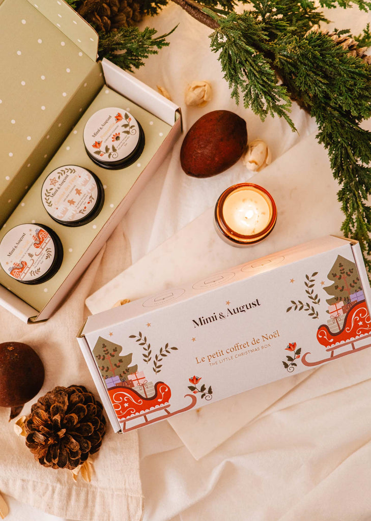 The Little Christmas Box by Mimi & August is a holiday gift set showcasing enchanting seasonal scents, featuring Christmas candles in festive packaging on a white surface, adorned with pine cones, nuts, and evergreen branches.