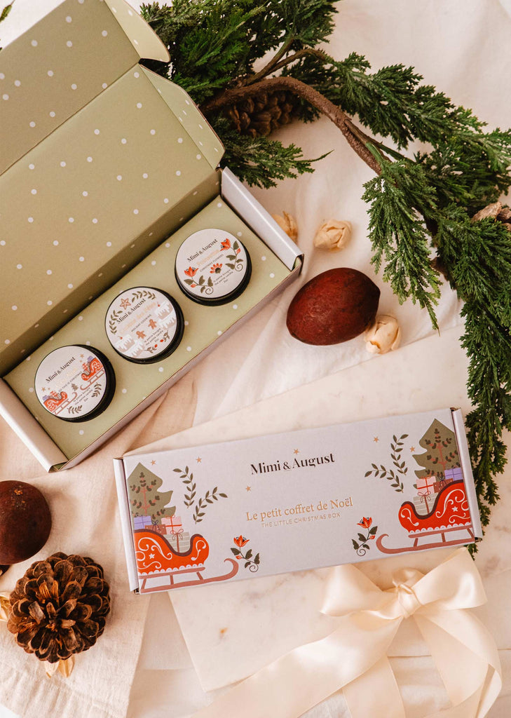 The Little Christmas Box by Mimi & August is a charming gift set containing three small jars elegantly presented in festive holiday packaging. Set against a backdrop of white fabric and adorned with pinecones, sprigs, and nuts, this collection embodies holiday-inspired fragrances ideal for any celebratory atmosphere.