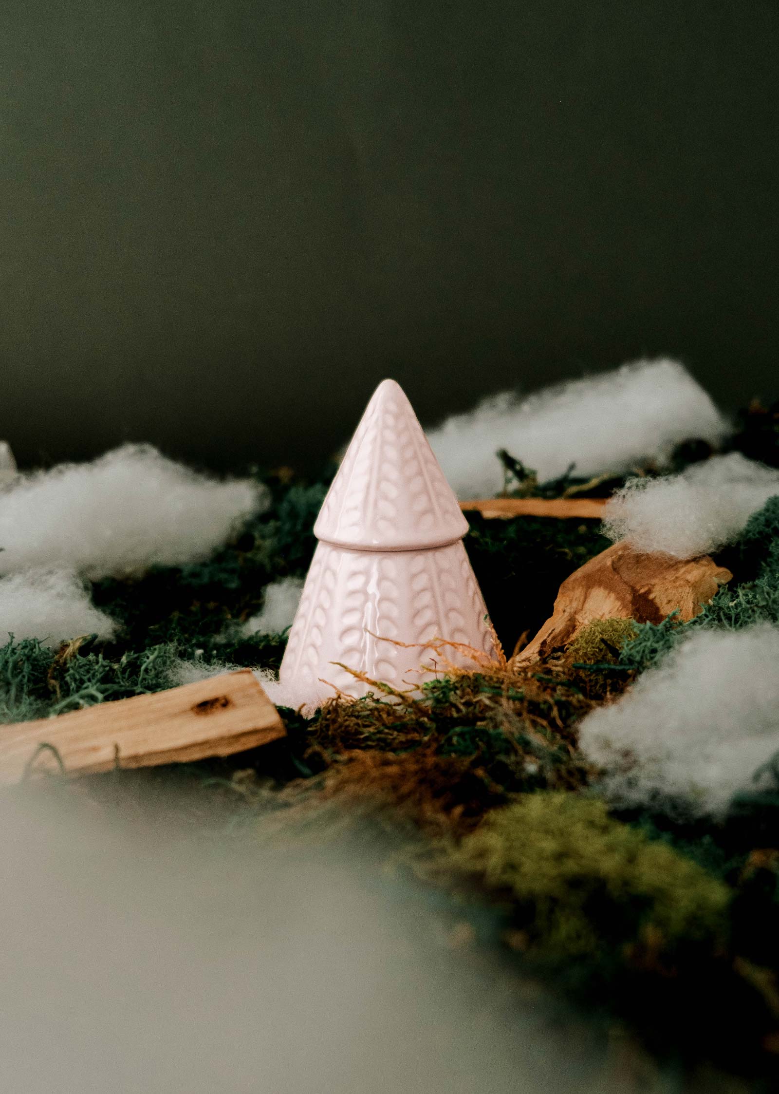 The Little Fir Tree by Mimi & August is a pink, cone-shaped decorative piece that looks like a Christmas candle, set among green moss and scattered wood pieces with fluffy white material surrounding it.