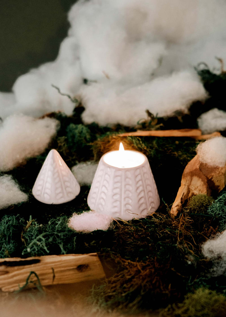 A burning "The Little Fir Tree" candle by Mimi & August and an unlit candle cover rest on moss with cotton pieces scattered around, radiating a festive aroma.