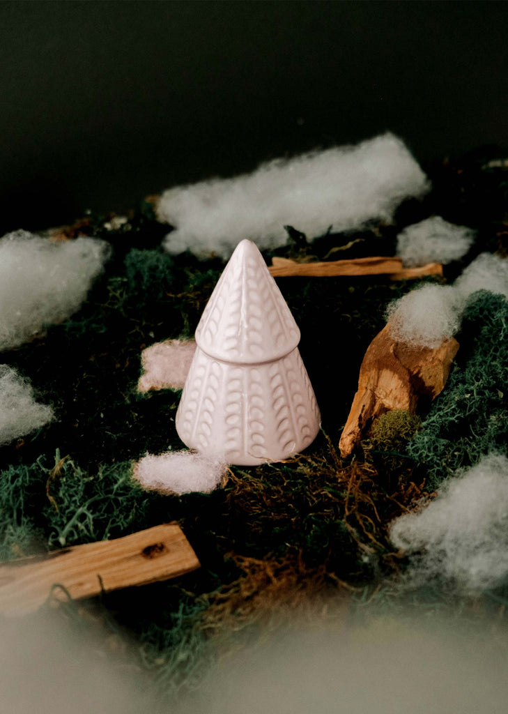 The Little Fir Tree by Mimi & August, a small pink ceramic figurine, sits on a textured surface alongside moss, wooden sticks, and white fluffy material, filling the air with a festive aroma.