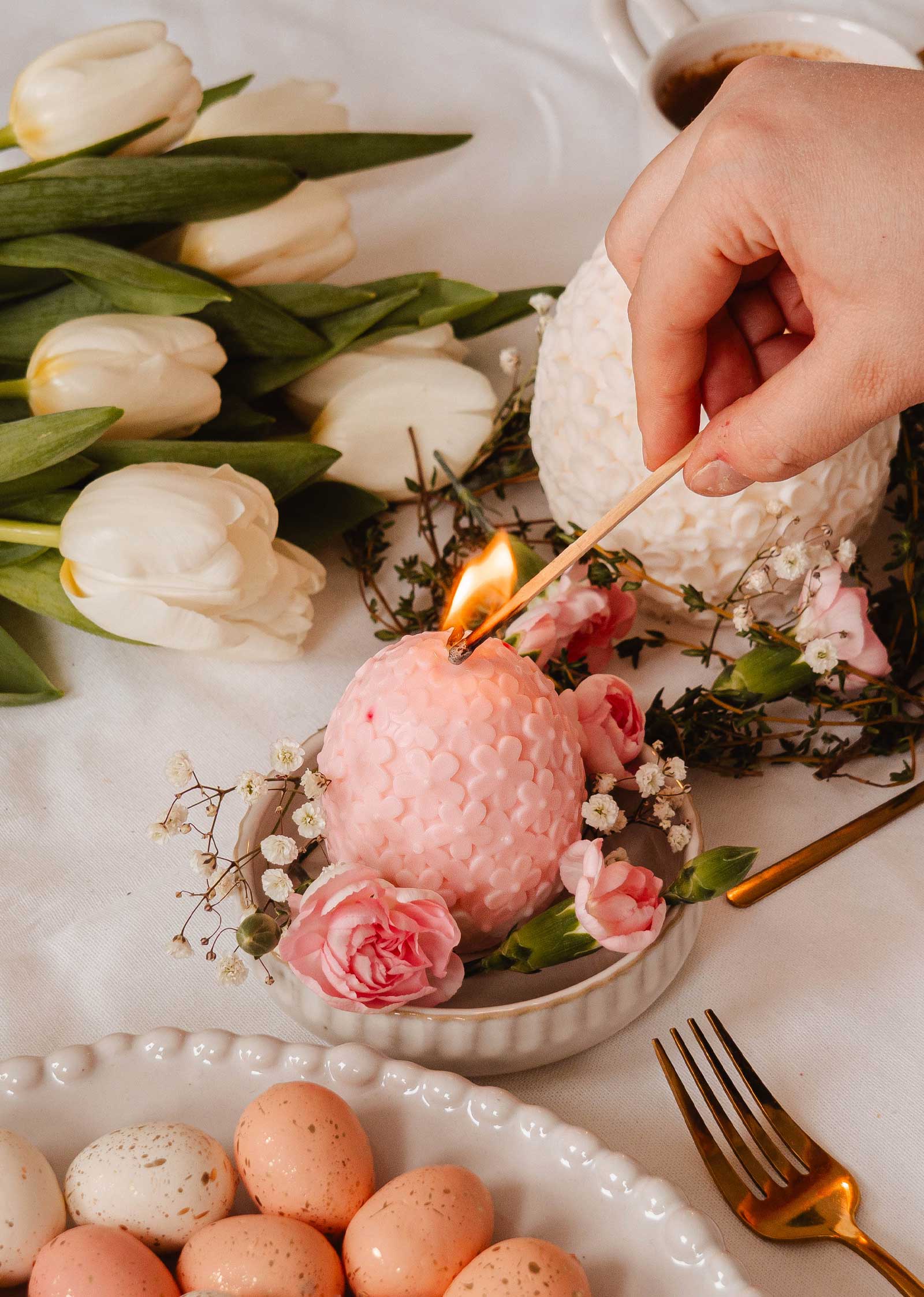 A hand lights The Little Floral Egg - Decorative Candle by Mimi & August, releasing a subtle floral scent amid blush pink roses and white tulips. Speckled eggs and a fork rest nearby, completing the elegant setting.