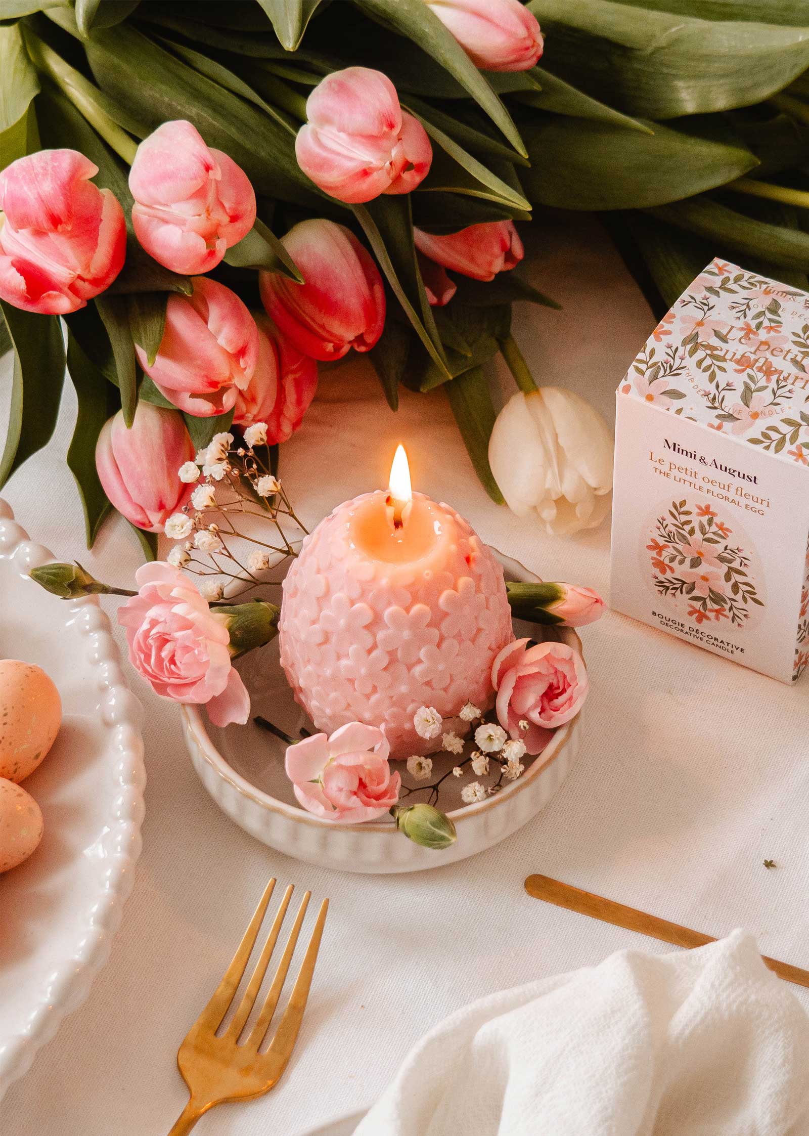 The Little Floral Egg by Mimi & August, a blush pink decorative candle adorned with tiny flowers, casts a soft floral aroma while flickering on the table. A gold fork gleams under its light, framed by pink tulips and a floral-patterned box for a serene atmosphere.