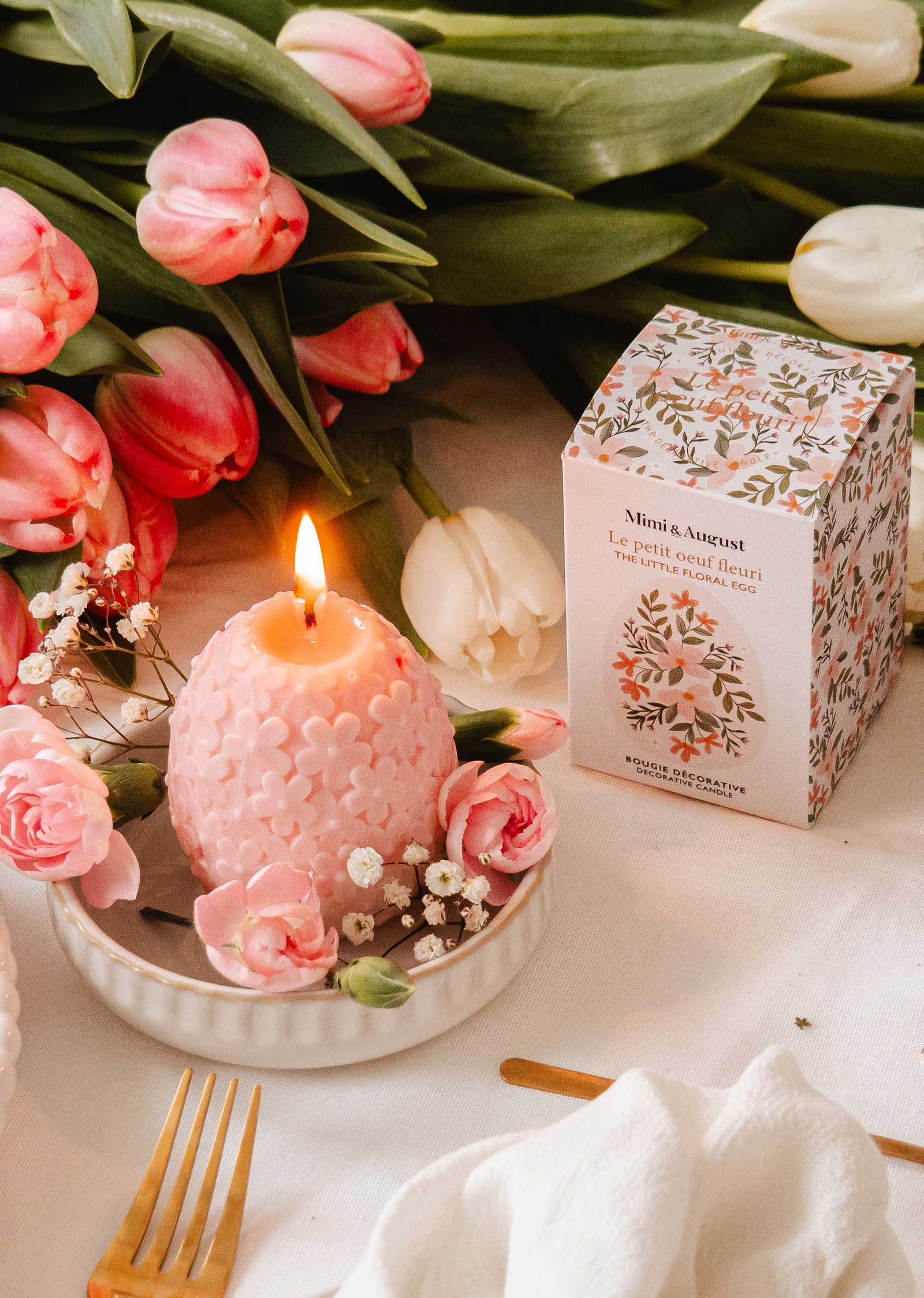 The Little Floral Egg - Decorative Candle by Mimi & August sits elegantly on a saucer, surrounded by small flowers. Its delightful floral scent fills the air, complemented by a decorative box and pink tulips on the white tablecloth.