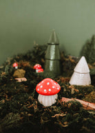 The Little Mushroom Candle by Mimi & August rests on moss, surrounded by other whimsical mushroom and tree décor, evoking an enchanted forest scene against a green backdrop.