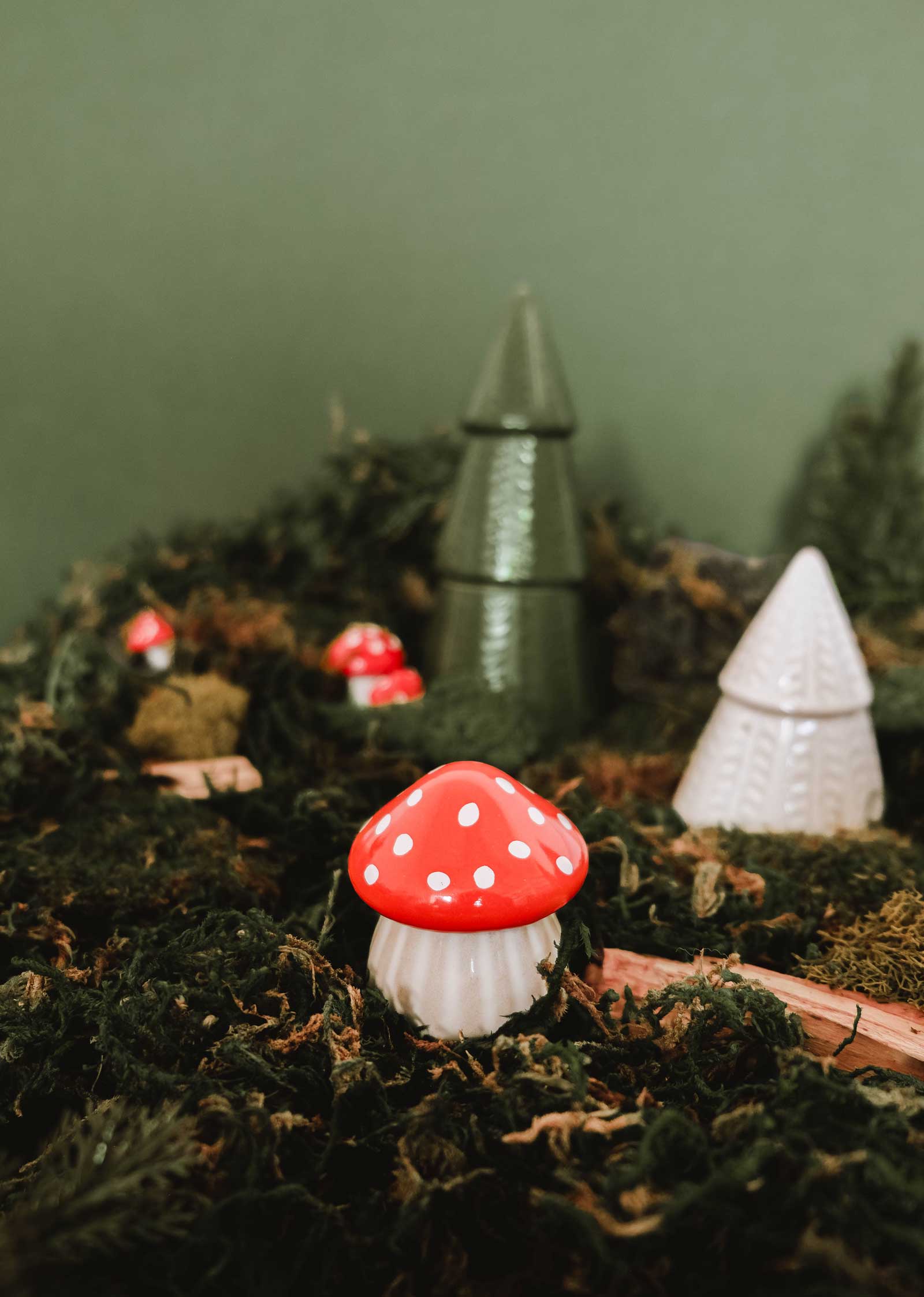 The Little Mushroom Candle by Mimi & August rests on moss, surrounded by other whimsical mushroom and tree décor, evoking an enchanted forest scene against a green backdrop.
