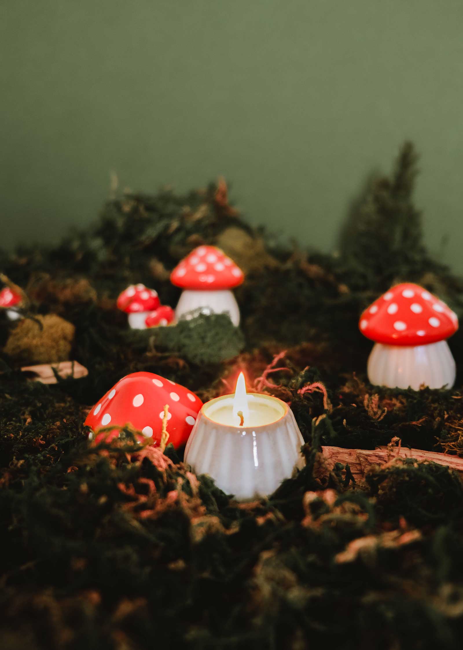 The Little Mushroom Candle by Mimi & August, in a white holder, is displayed on moss with faux red and white mushrooms and a green background.