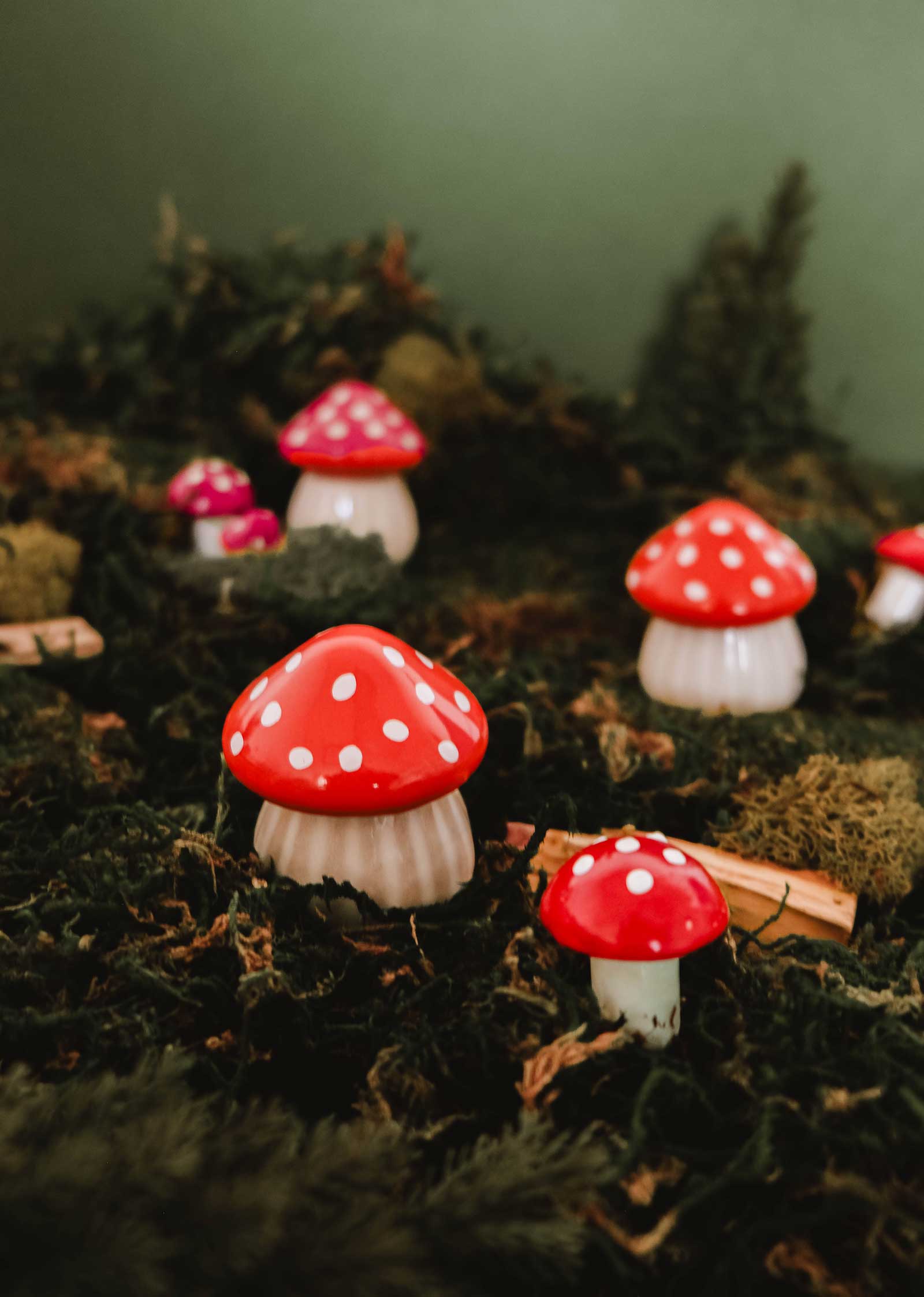 Several red and white spotted mushrooms, resembling The Little Mushroom Candle by Mimi & August, grow from moss-covered ground with a green background and scattered pieces of wood.