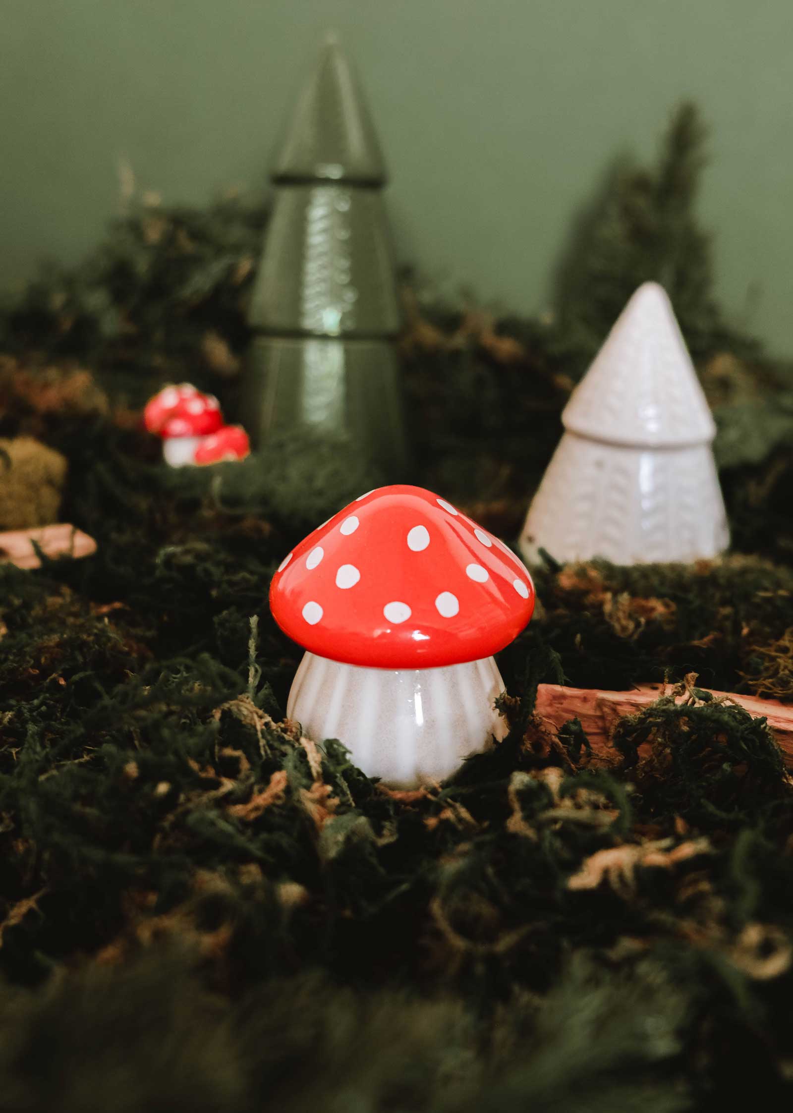 A ceramic red and white mushroom figurine sits among green moss, with cone-shaped tree ornaments and The Little Mushroom Candle by Mimi & August charmingly displayed in the background.