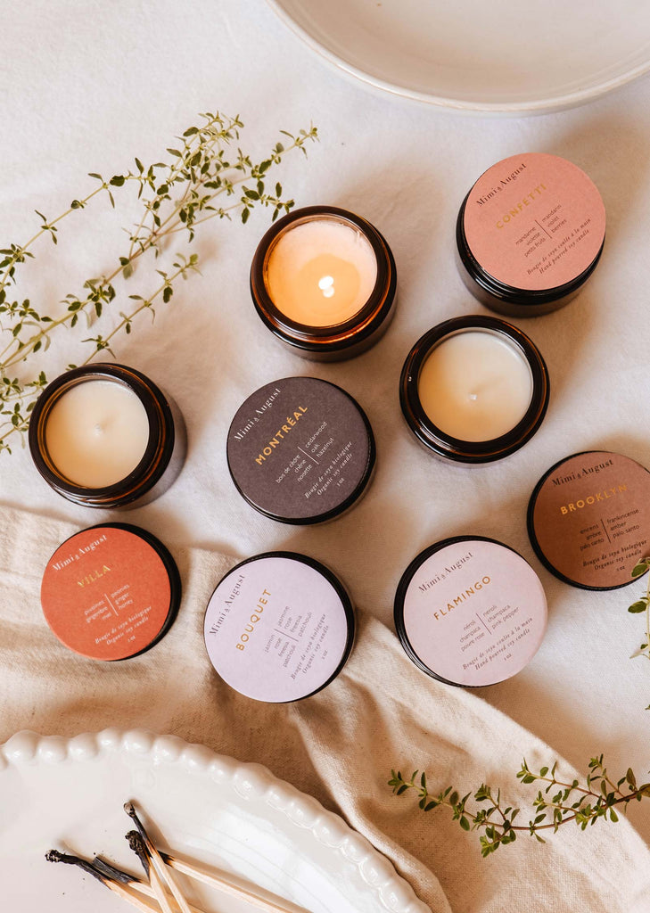 A variety of scented candles in amber jars with colorful lids from The Nostalgia Bundle by Mimi & August are arranged on a white cloth, accompanied by green sprigs and wooden matchsticks for a delightful surprise scent experience.