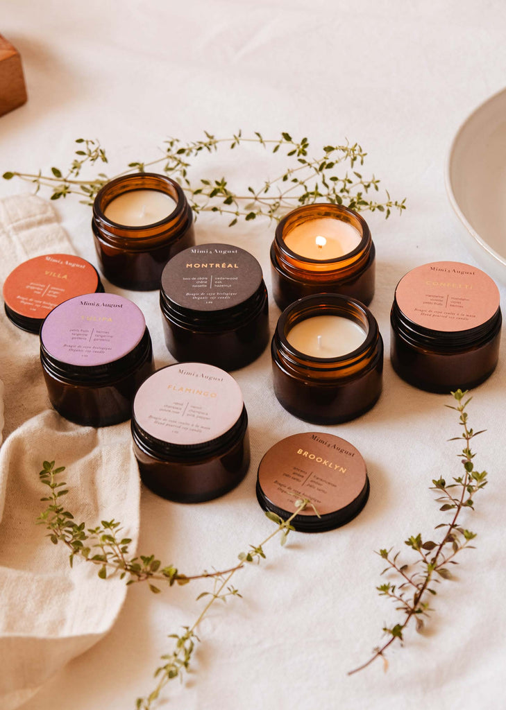 An assortment of nine small, amber glass candles, each with a unique labeled lid and surrounded by green sprigs on a beige fabric surface, creates the perfect Nostalgia Bundle from Mimi & August.