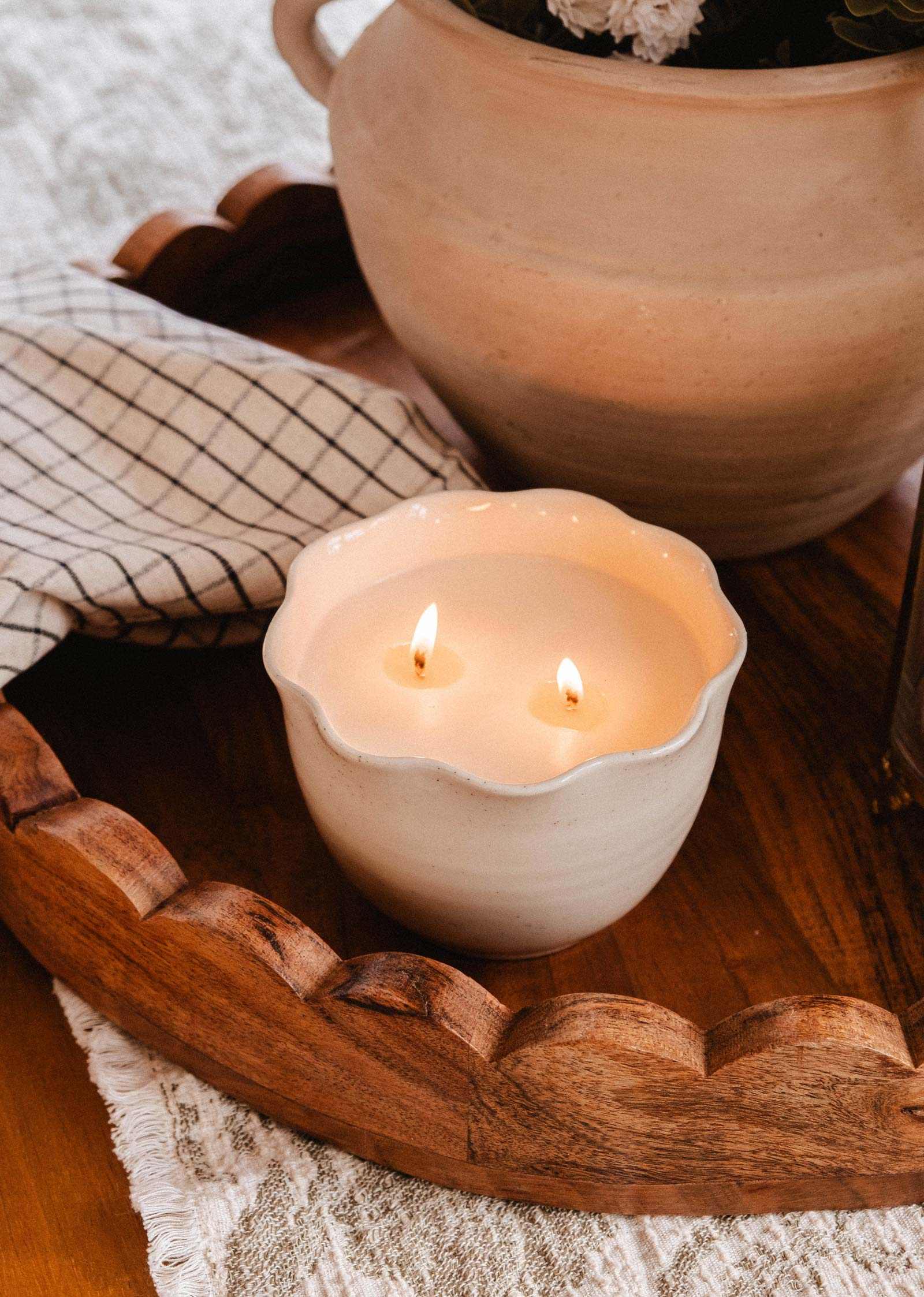 The Petal Bowl Candle by Mimi & August features two lit wicks in a handcrafted ceramic bowl, set on a wooden tray beside a checkered cloth and beige pot—an ideal Mother's Day gift.