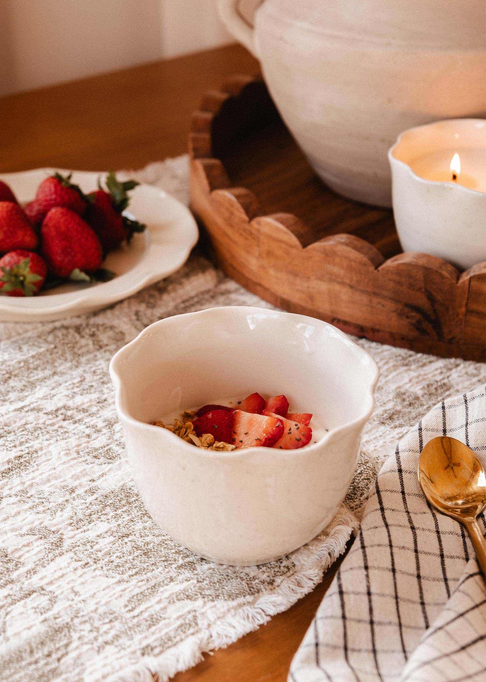A white bowl with yogurt, granola, and strawberries rests on a textured cloth beside a gold spoon and checkered napkin; in the background, fresh strawberries and Mimi & August's The Petal Bowl Candle add a soft glow.
