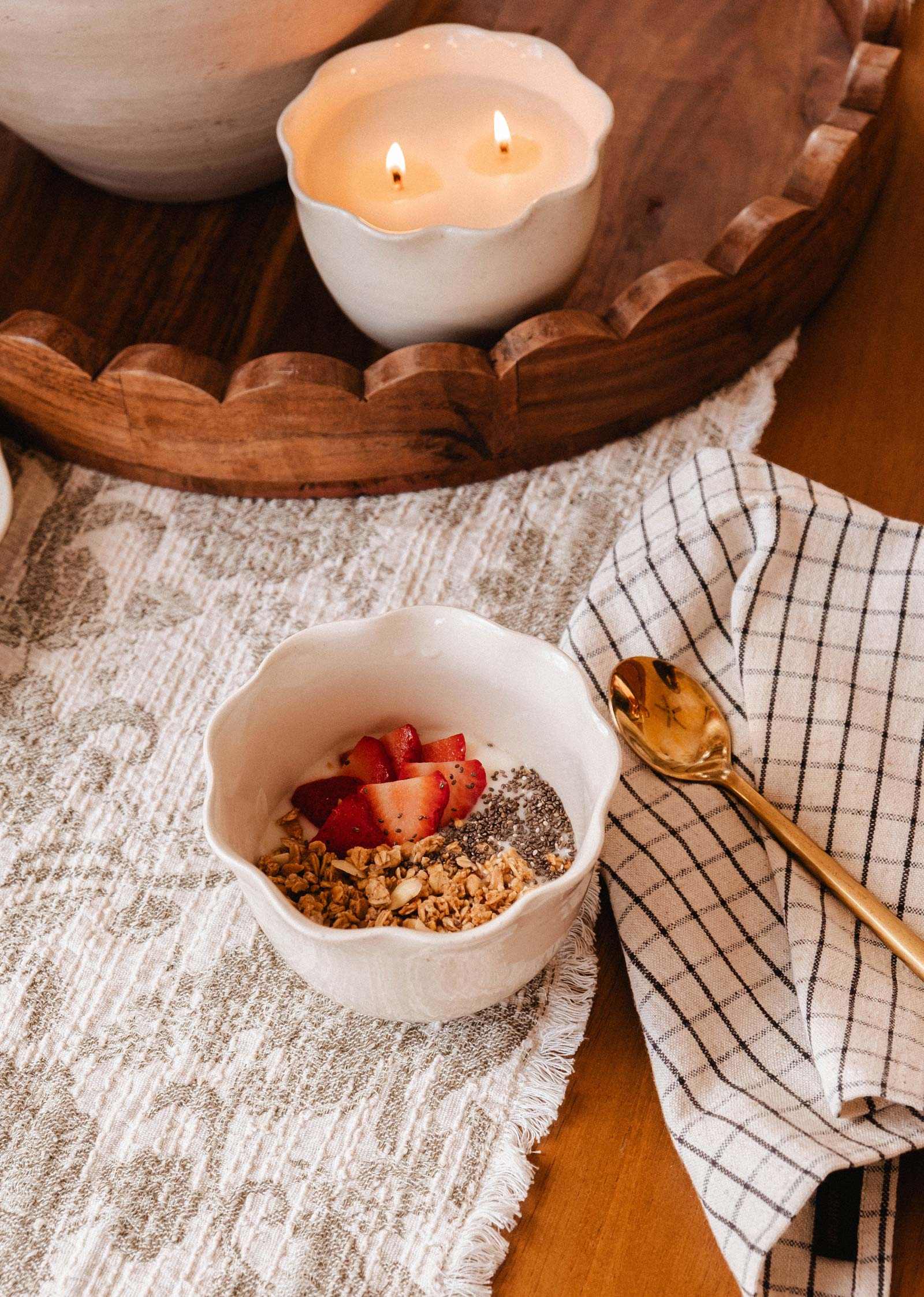 A bowl of yogurt with granola, chia seeds, and strawberries sits by a gold spoon, checkered napkin, and Mimi & August’s The Petal Bowl Candle—a lovely limited edition piece perfect for Mother’s Day—in the background.