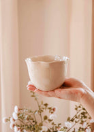 A hand holds The Petal Bowl Candle by Mimi & August, a light beige, scalloped-edge handcrafted ceramic candle with a glossy finish—ideal as a limited edition piece or thoughtful Mother's Day gift; blurred flowers in the background.