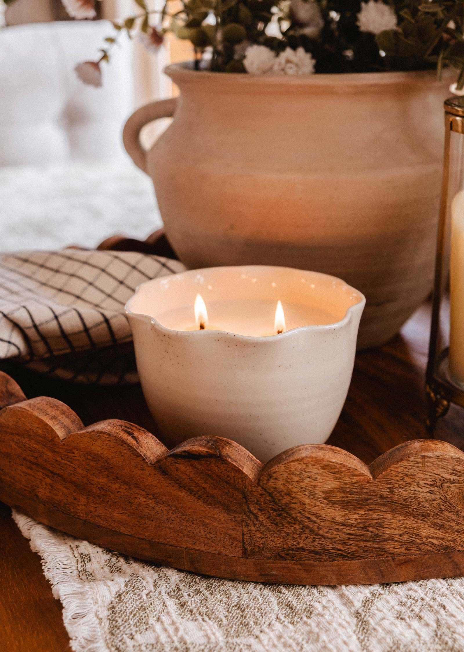 The Petal Bowl Candle by Mimi & August features two flames in a white ceramic holder. Set on a lace tablecloth beside a wooden tray, checked napkin, and beige floral vase, it makes a perfect Mother's Day gift.