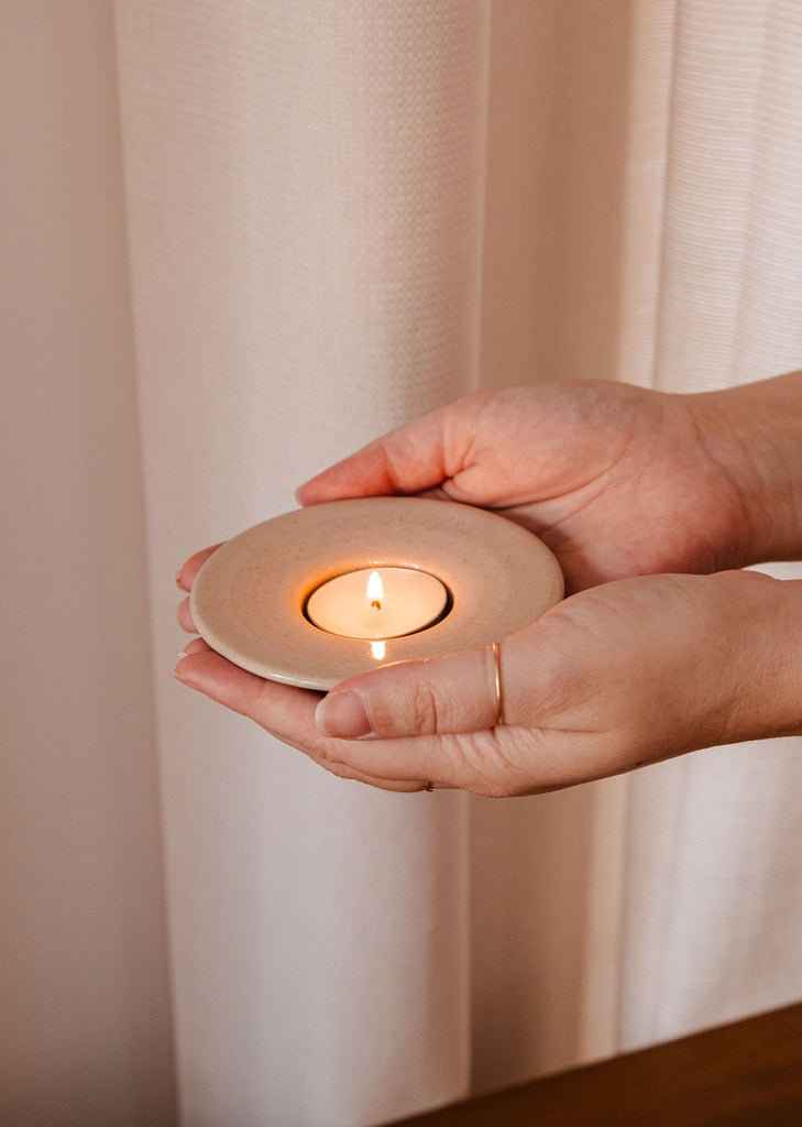 A person's hand holding a Mimi & August Round Tealight Holder with a candle on a plate, exuding rustic charm.