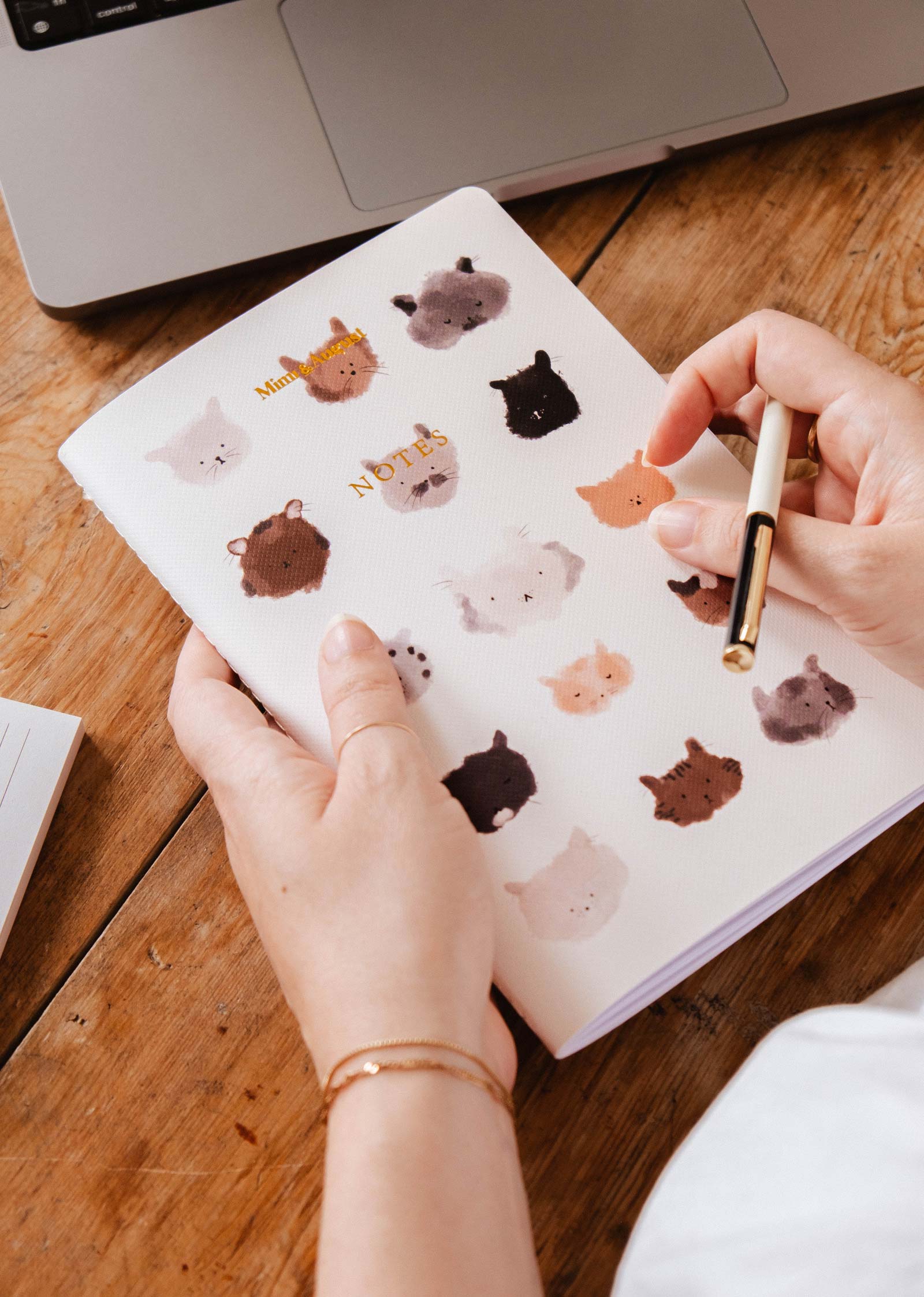 A person holds a pen and The Purrfect - Notebook by Mimi & August, featuring illustrated animal faces, next to a laptop on a wooden table.