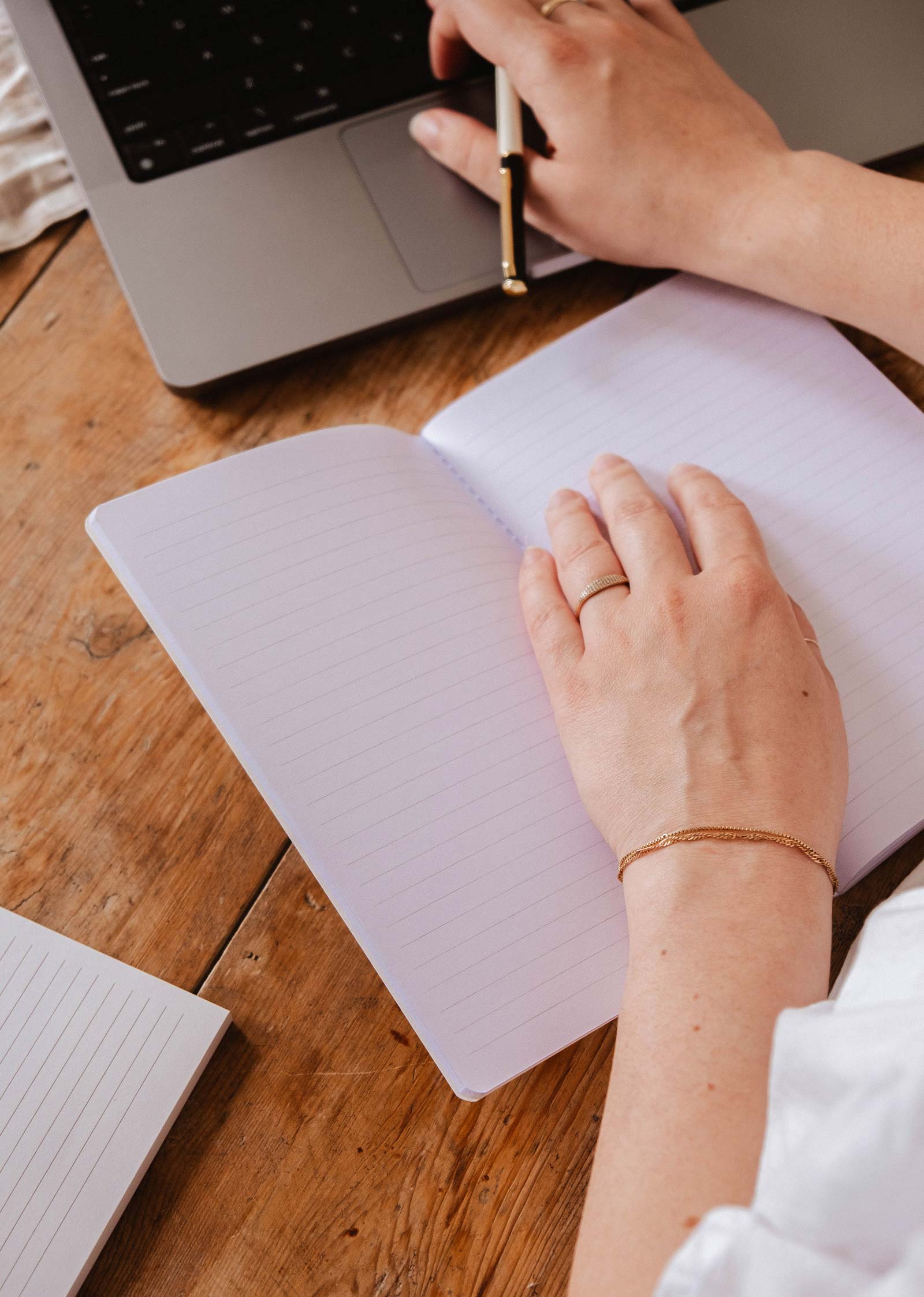 Someone writes in The Purrfect - Notebook by Mimi & August, with cute stationery next to an open laptop on a wooden desk.