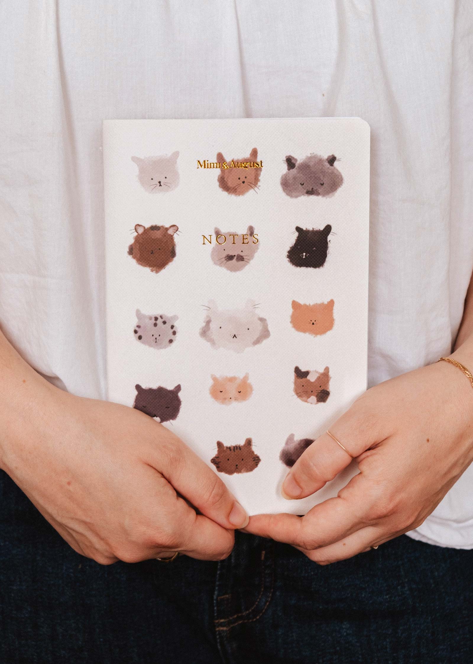 Someone holds The Purrfect - Notebook by Mimi & August, a white notebook with illustrated animal faces in a grid on the cover—ideal for cute stationery lovers.