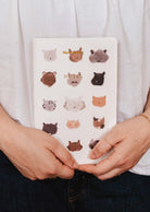 Someone holds The Purrfect - Notebook by Mimi & August, a white notebook with illustrated animal faces in a grid on the cover—ideal for cute stationery lovers.