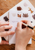 Someone writes on the Mimi & August Purrfect - Notepad, adorned with illustrated kitty faces, using a black and gold pen on a wooden surface.