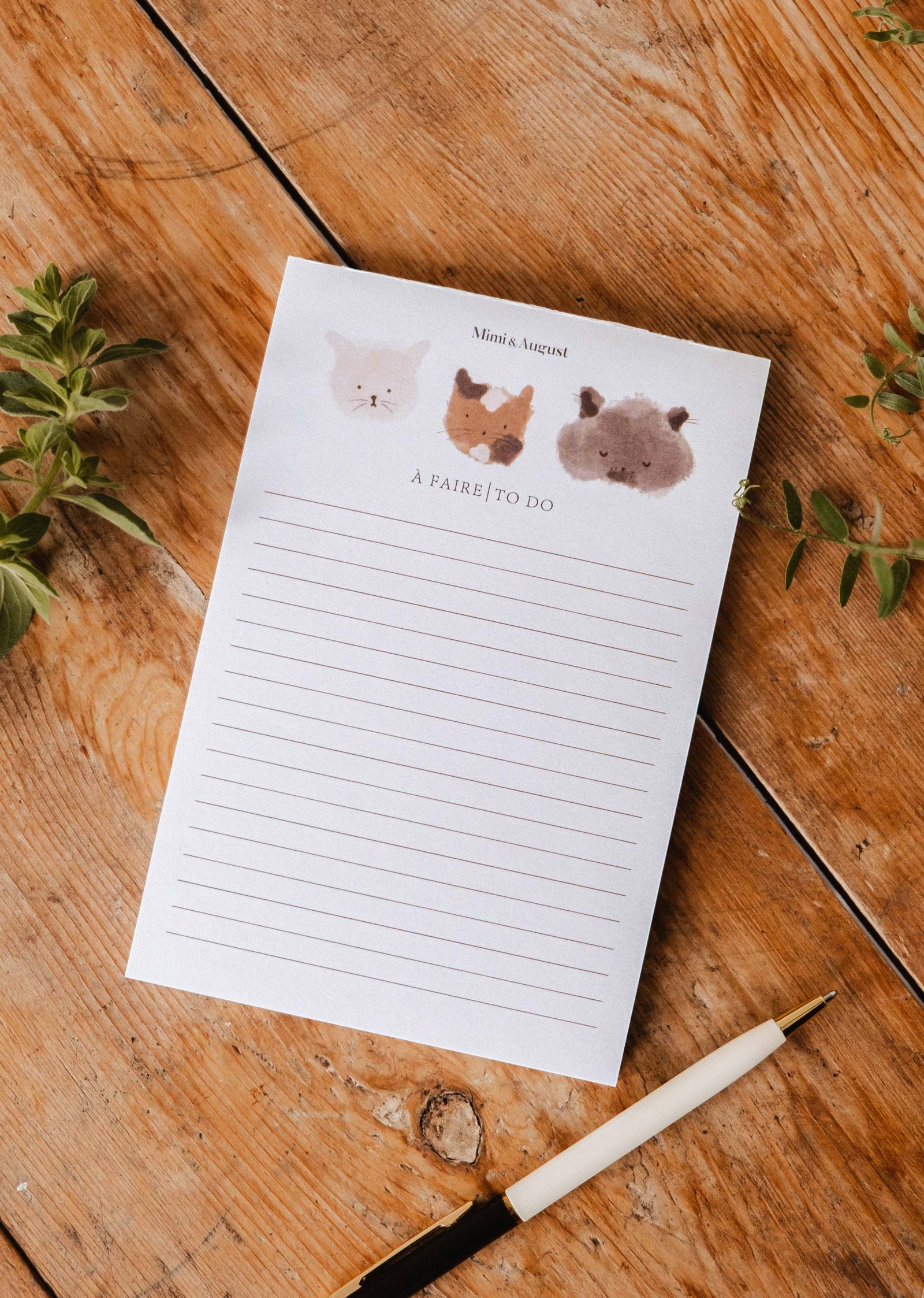 The "Purrfect - Notepad" by Mimi & August features adorable kitty illustrations at the top and lined pages, perfect for jotting down to-dos. Shown on a wooden surface beside a pen and green plants.