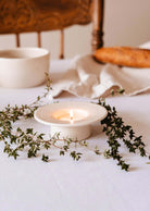 A lit candle in The Round Tealight Holder by Mimi & August sits on a white tablecloth, surrounded by green foliage, with a loaf of bread and bowl enhancing the serene scene.