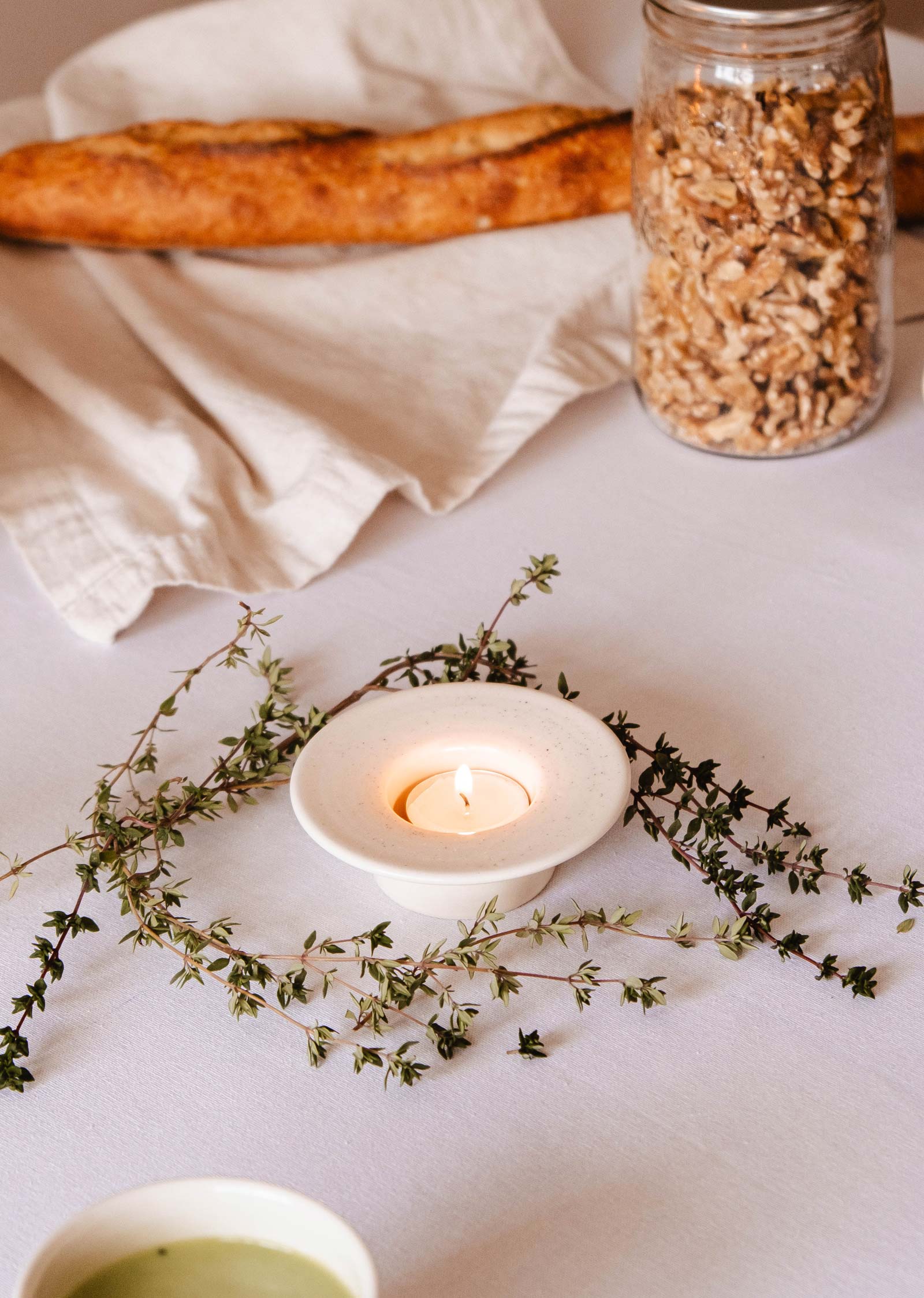 Une bougie allumée dans "The Round Tealight Holder" de Mimi & August est posée sur une table blanche entourée de branches de thym, avec un pot de noix et une baguette en arrière-plan.