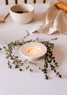 The Round Tealight Holder by Mimi & August cradles a lit candle amid thyme sprigs on a white tablecloth, with a bowl and napkin nearby.