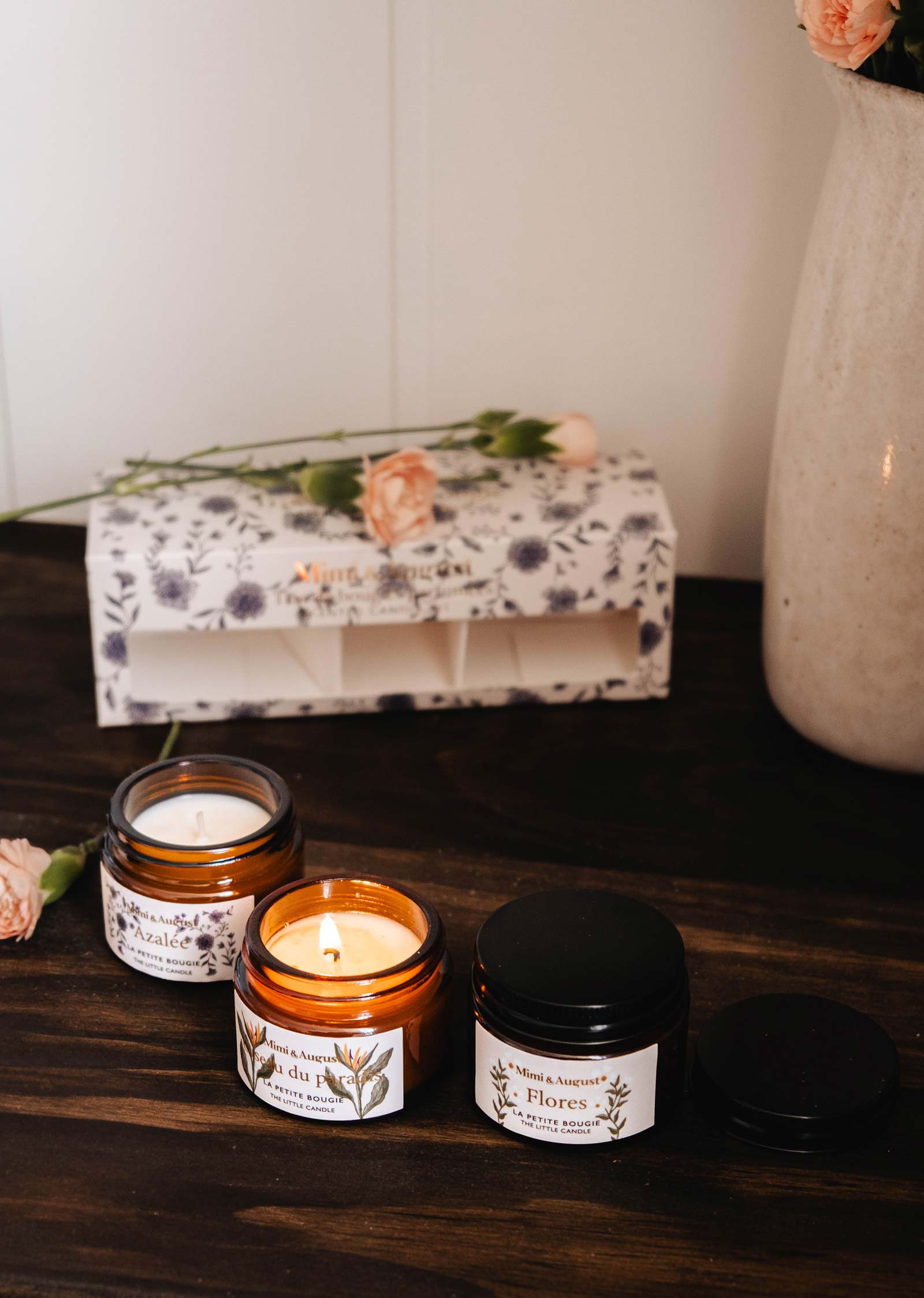 Three small floral-scented candles with delicate labels rest on a wooden surface, one lit. Accompanied by the Mimi & August Secret Garden Box Set and a vase of peach flowers, the setting exudes serenity.