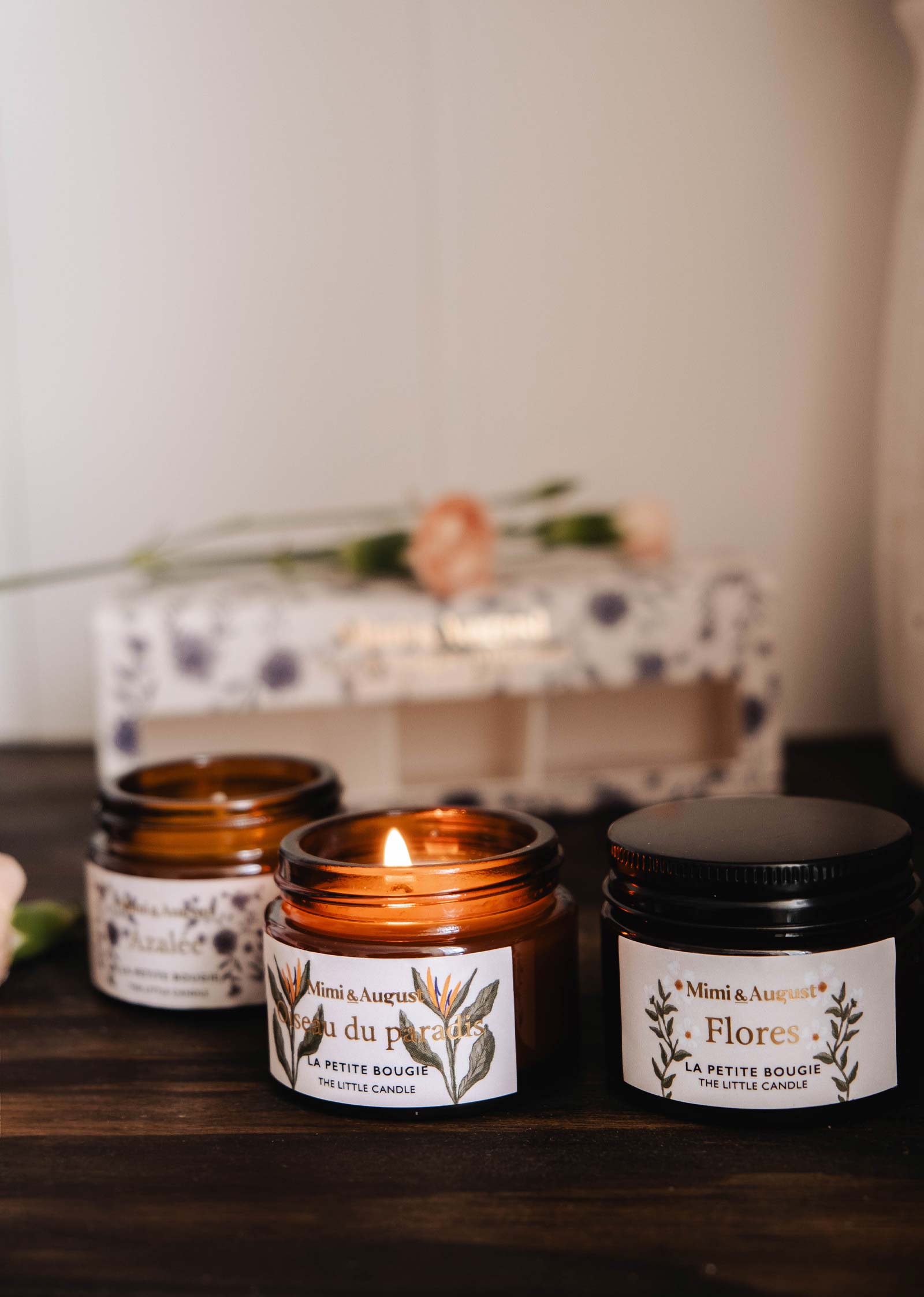 Three small amber glass candles with botanical labels from Mimi & August's The Secret Garden Box Set are on a wooden surface, one lit, with a blurred floral box in the background.